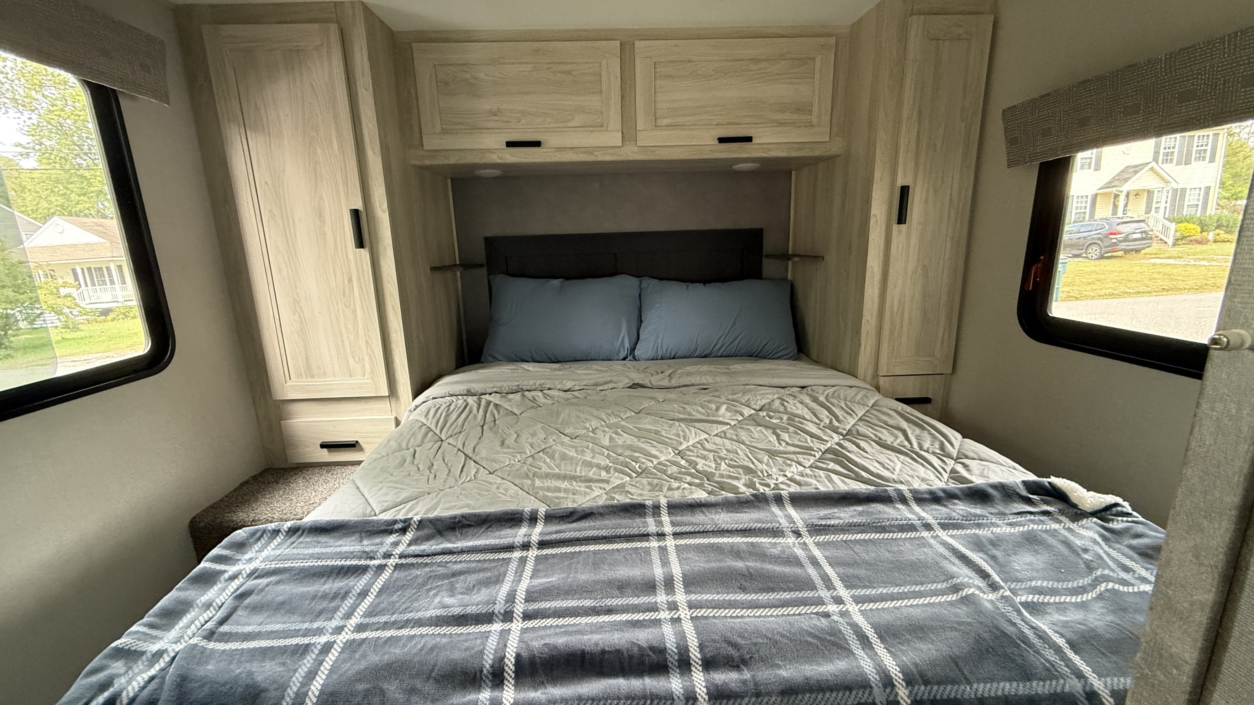 Comfortable RV rental bedroom with a bed, blue pillows, plaid blanket, wood cabinets, and outside views through the windows.. Forest River Sunseeker LE 2023