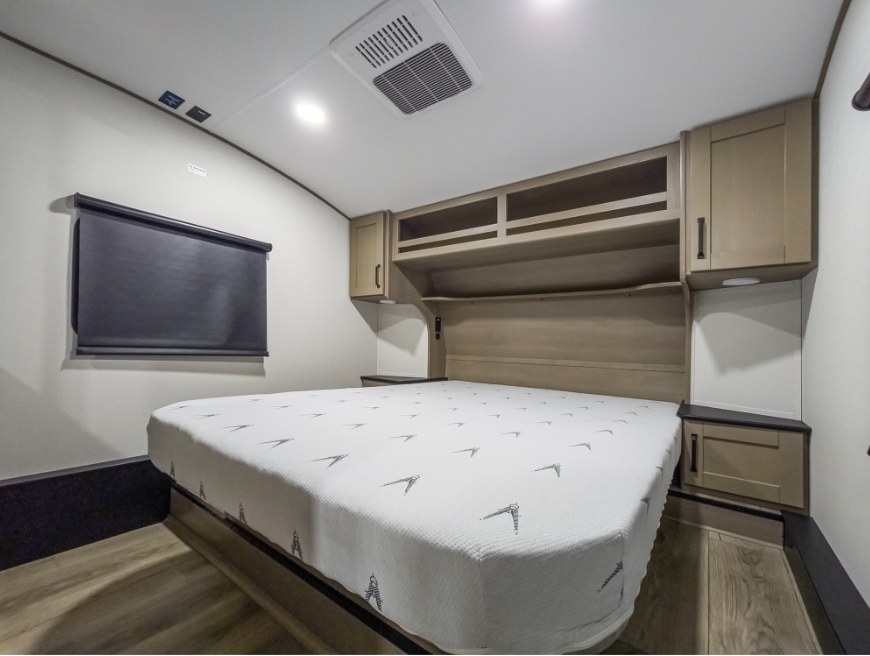 RV rental bedroom featuring a modern bed with patterned mattress, wood-toned cabinets, and a dark window blind.. Grand Design Reflection 100 2026