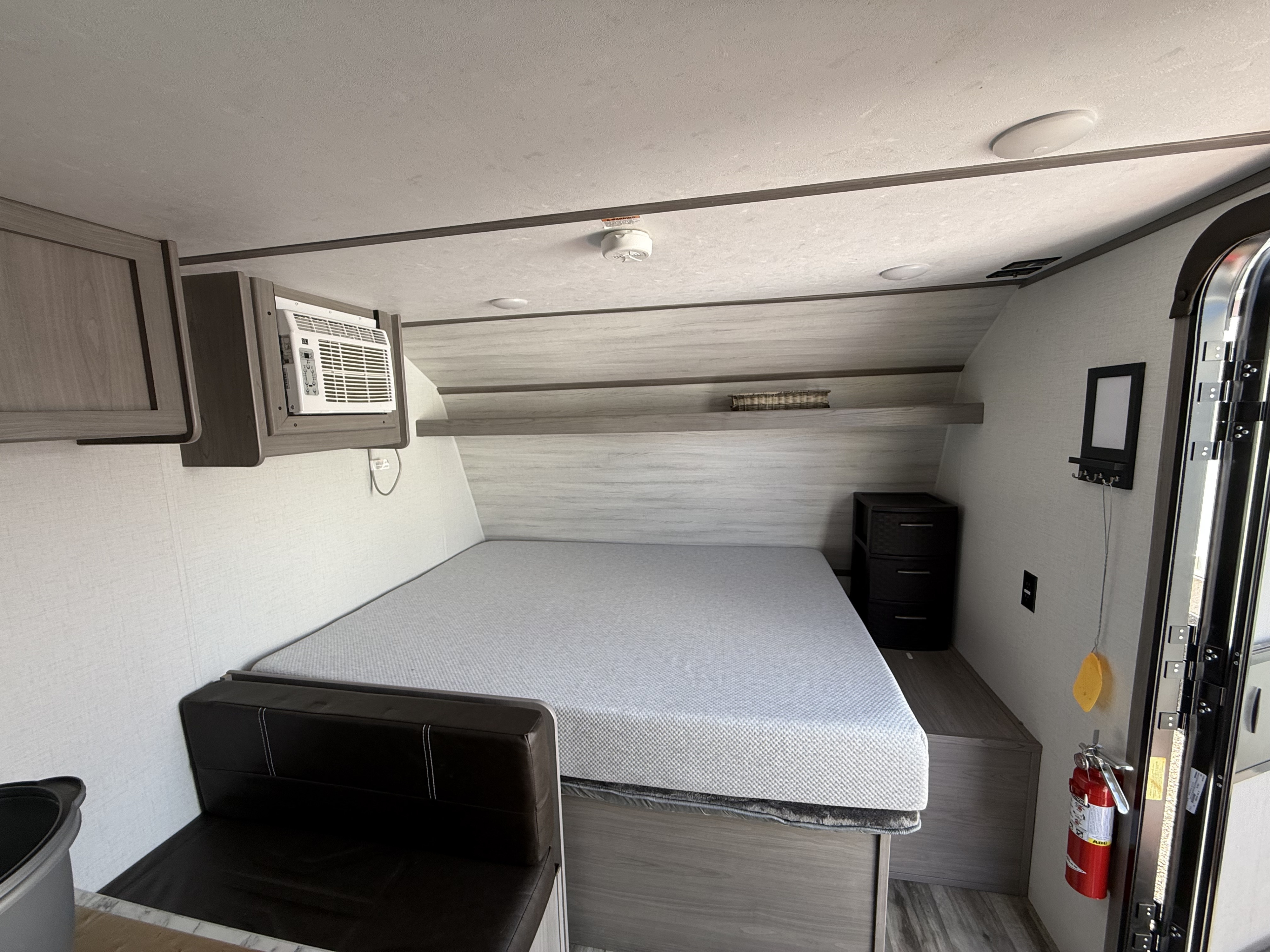 RV rental interior featuring a cozy bed, AC unit, overhead cabinets, and a fire extinguisher by the door for safety.. Dutchmen Coleman Lantern LT 2023