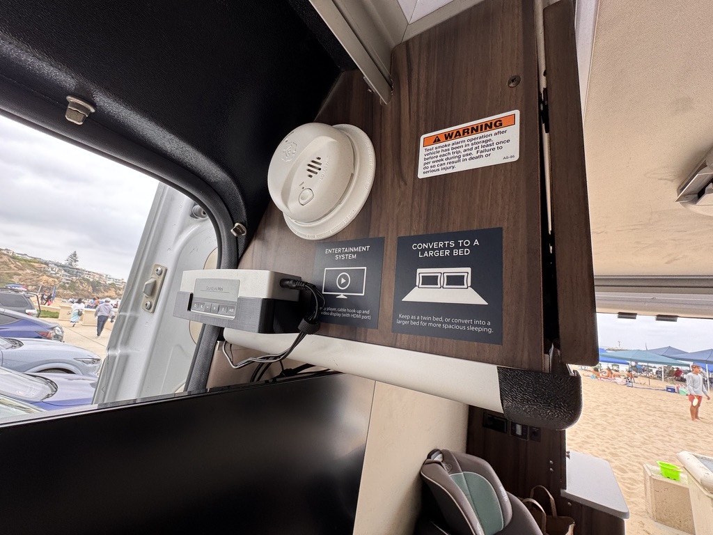 RV rental interior: smoke alarm, retro entertainment console, and bed conversion guide. Outside, a lively beach view.. Carado Carado Class B 2019