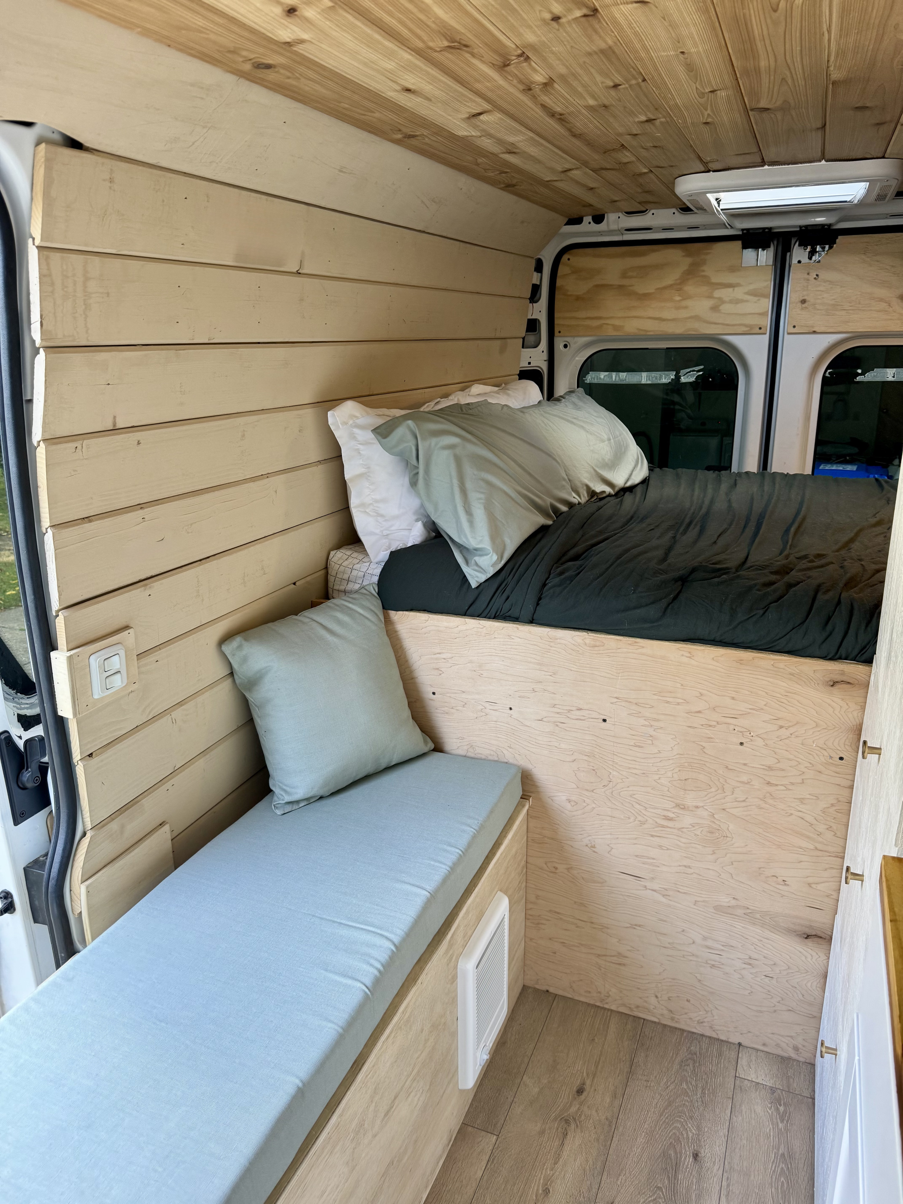 Warm RV rental interior with wood plank walls & ceiling, a dark bed, and light blue cushioned bench for travel.. Dodge Ram Pro Master 2023