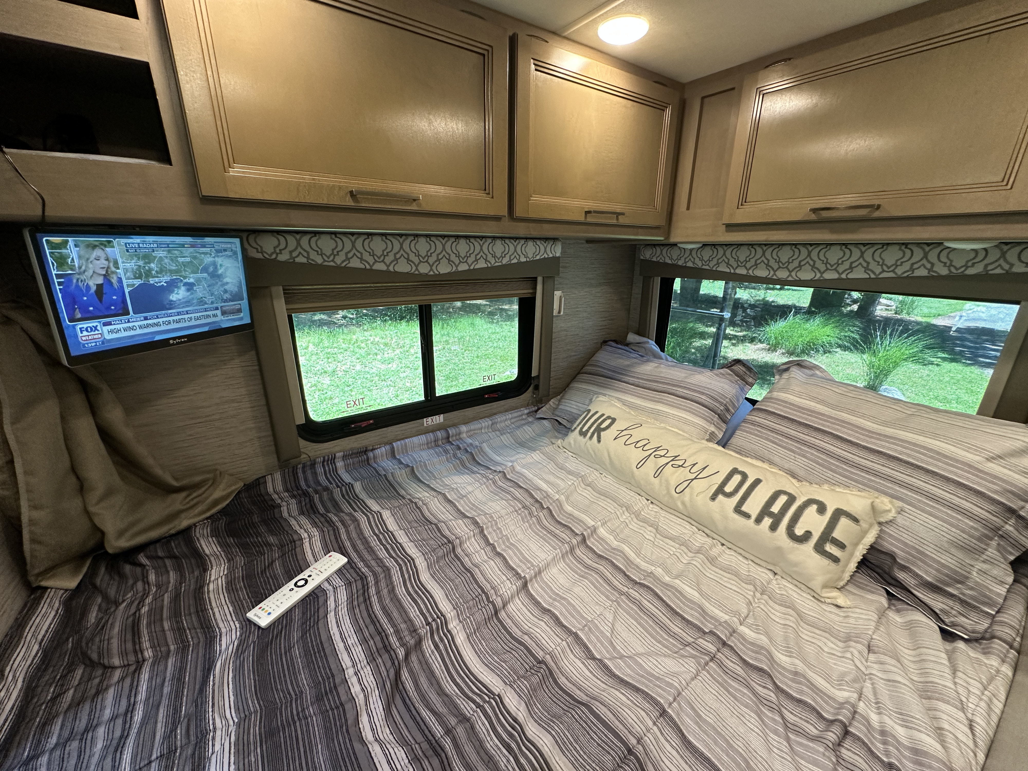 Cozy RV rental bedroom with striped bedding, a "happy PLACE" pillow, and a TV showing weather for comfortable travel.. Thor Quantum SE 2021