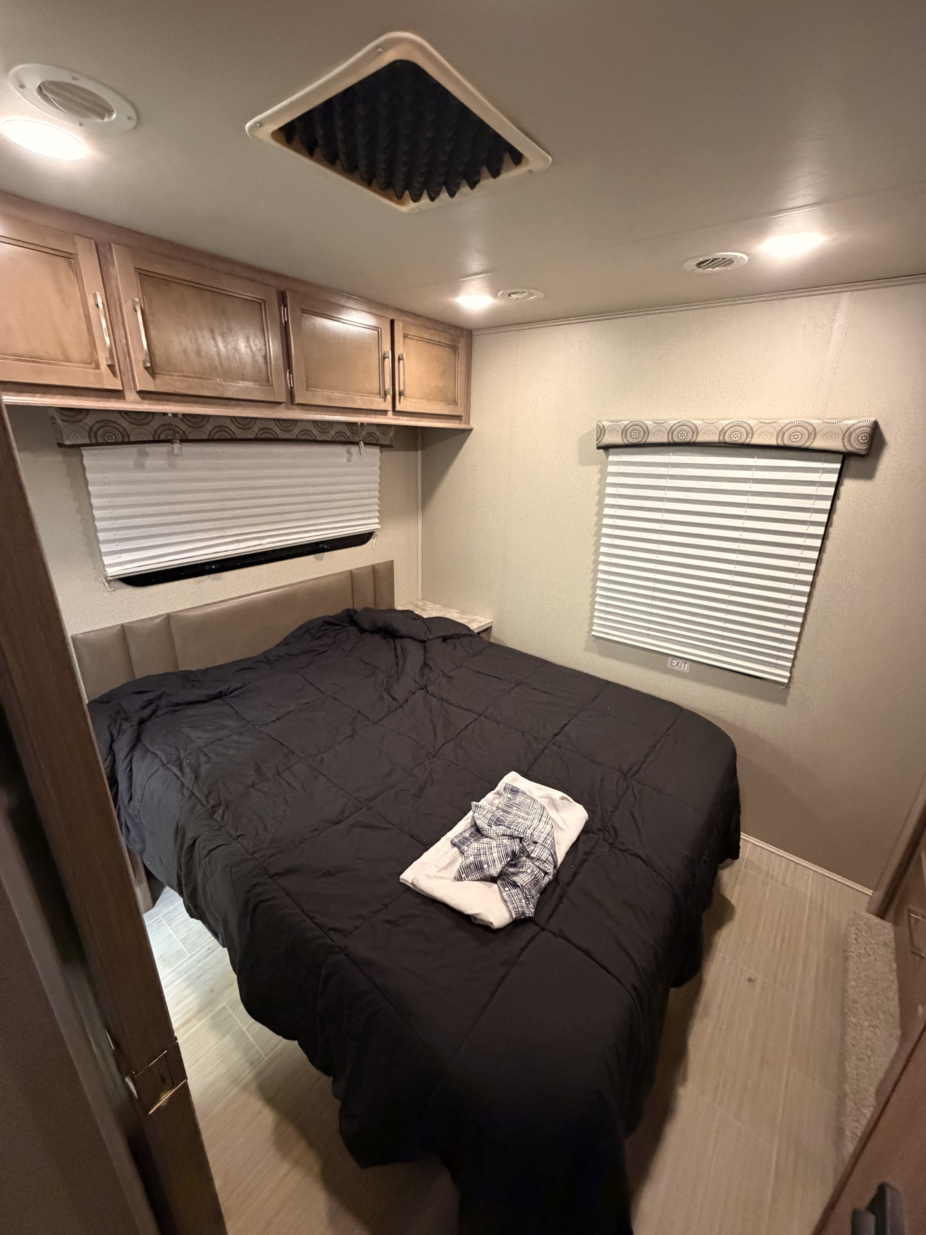Inside this RV rental, a compact bedroom showcases a bed with black comforter, overhead cabinets, and window blinds.. Entegra Coach Odyssey 2020