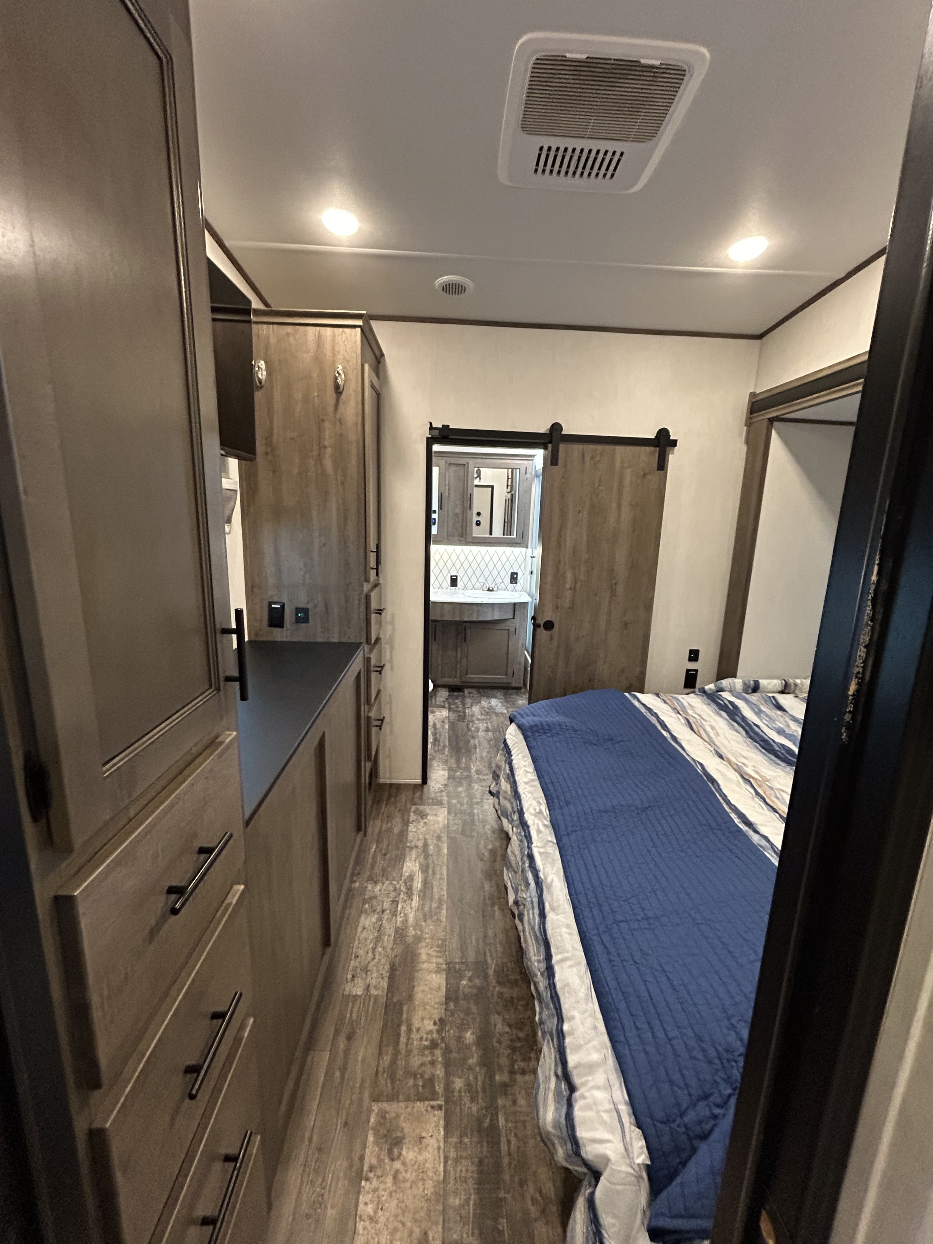 This RV rental bedroom features ample wood storage, plank flooring, a bed with blue bedding, and a private bathroom through a barn door.. Forest River Sabre 2022