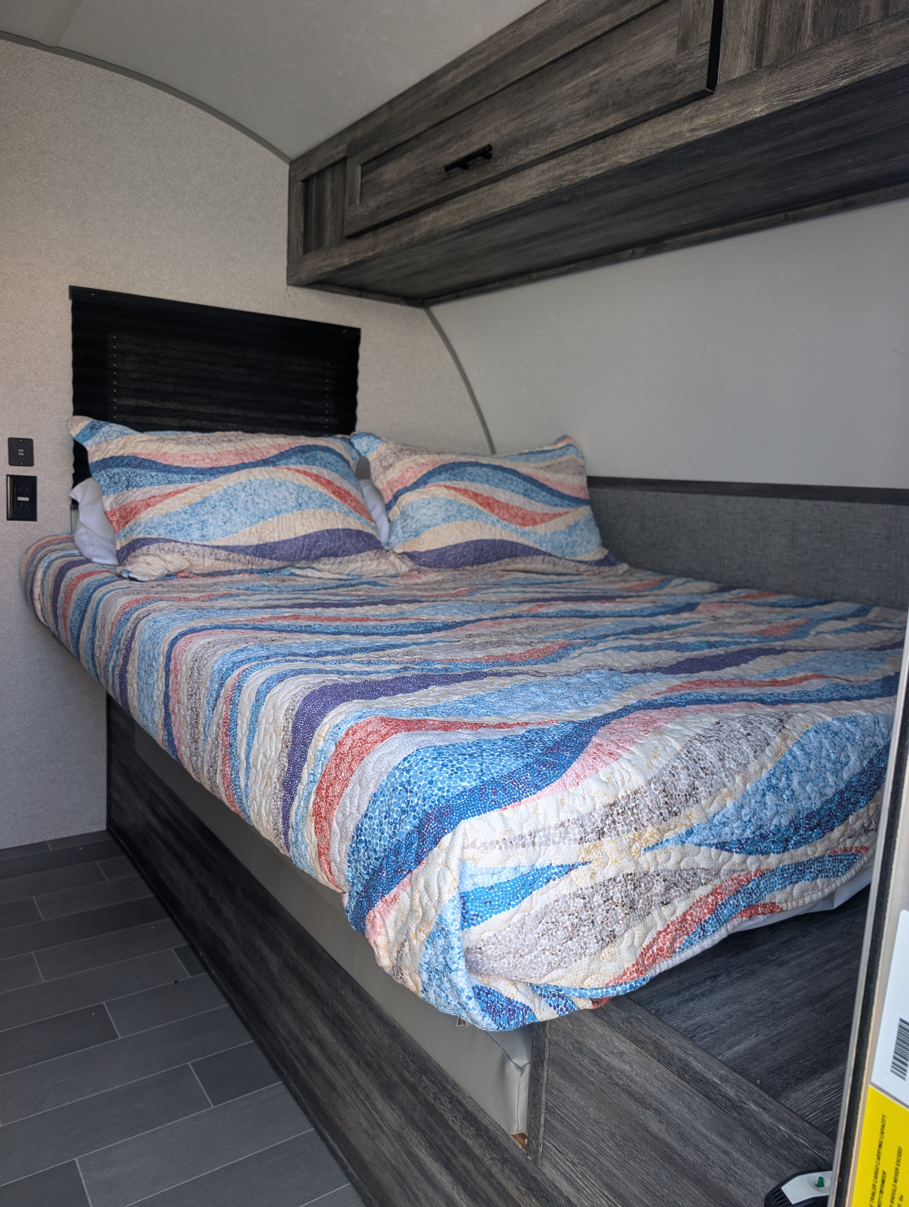 Cozy RV rental bedroom featuring a bed with a colorful wavy quilt, pillows, overhead storage, window blind, and dark tile floor.. Forest River Wildwood FSX Plantium 2022