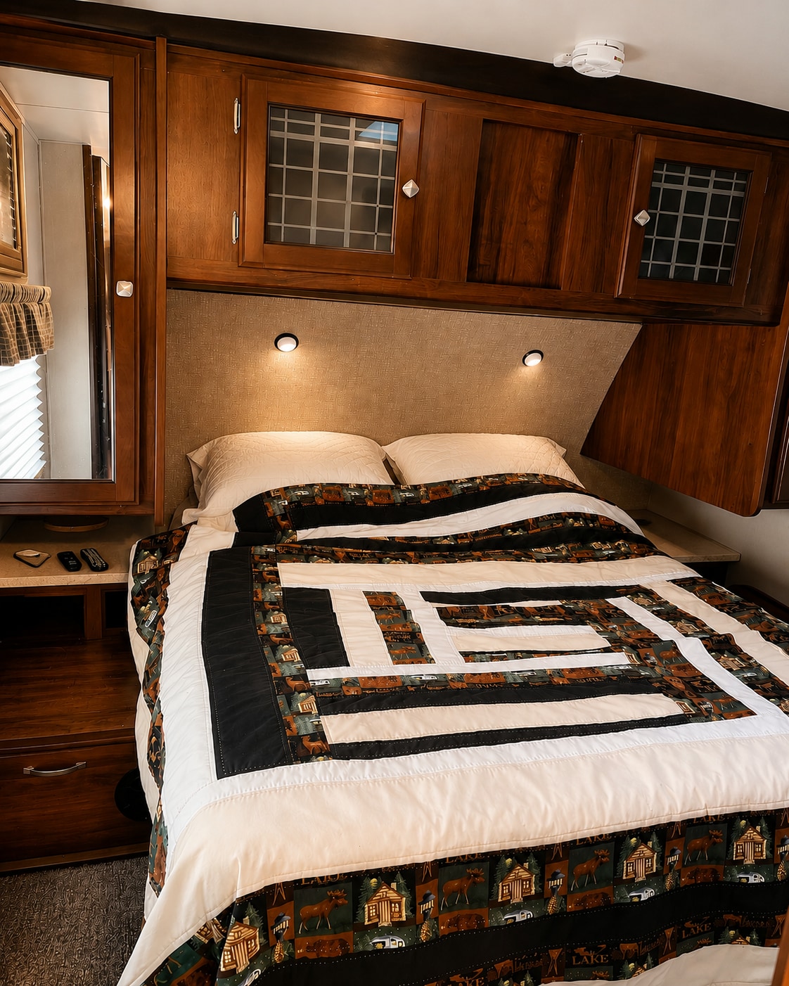 Cozy RV rental bedroom featuring a bed with a rustic-themed quilt, illuminated headboard, and overhead storage.. Heartland RVs Mallard 2019