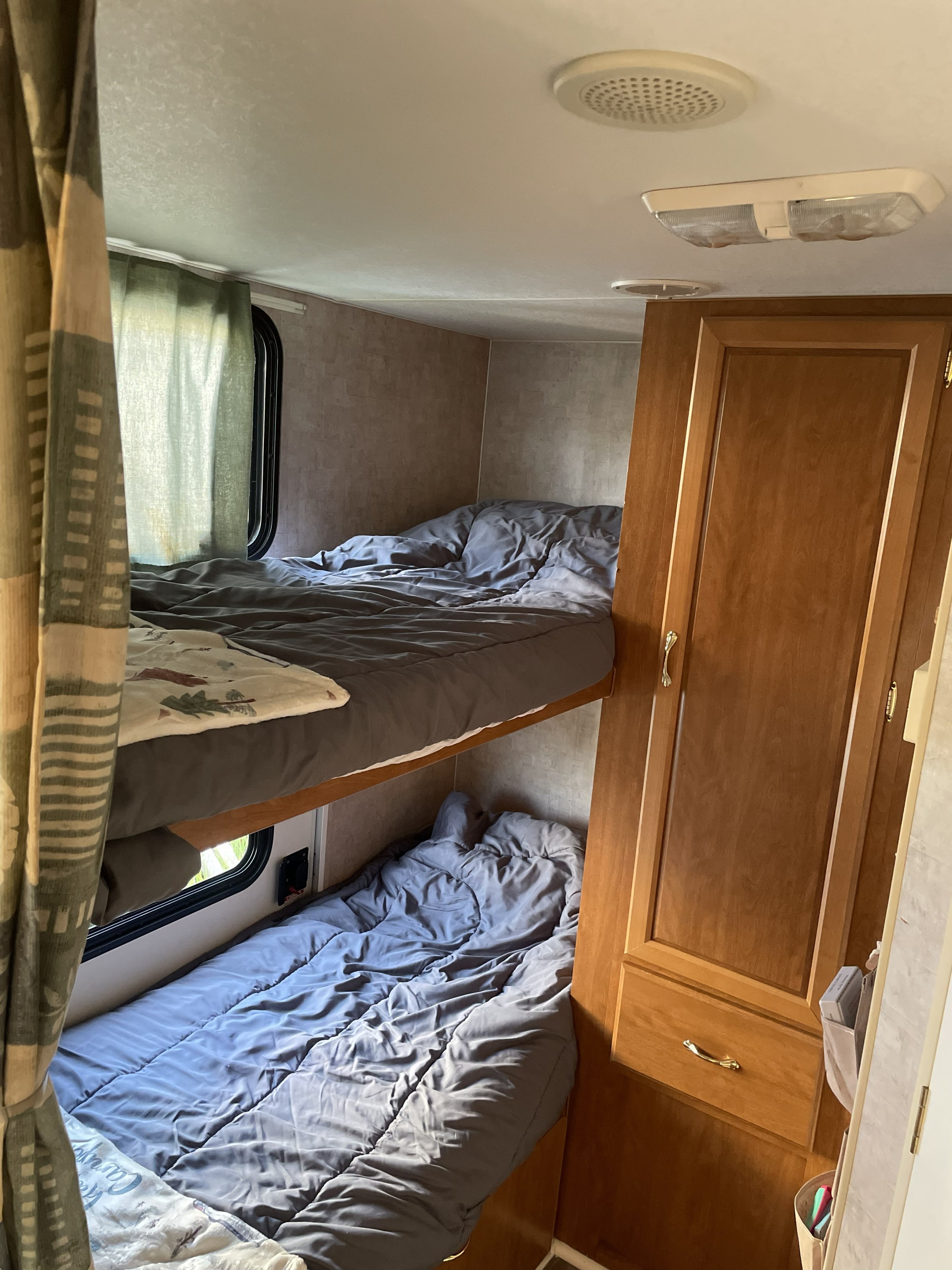Cozy bunk beds in an RV rental, featuring plush grey bedding, a window with curtains, and integrated wooden cabinetry.. Fleetwood Mallard 2005