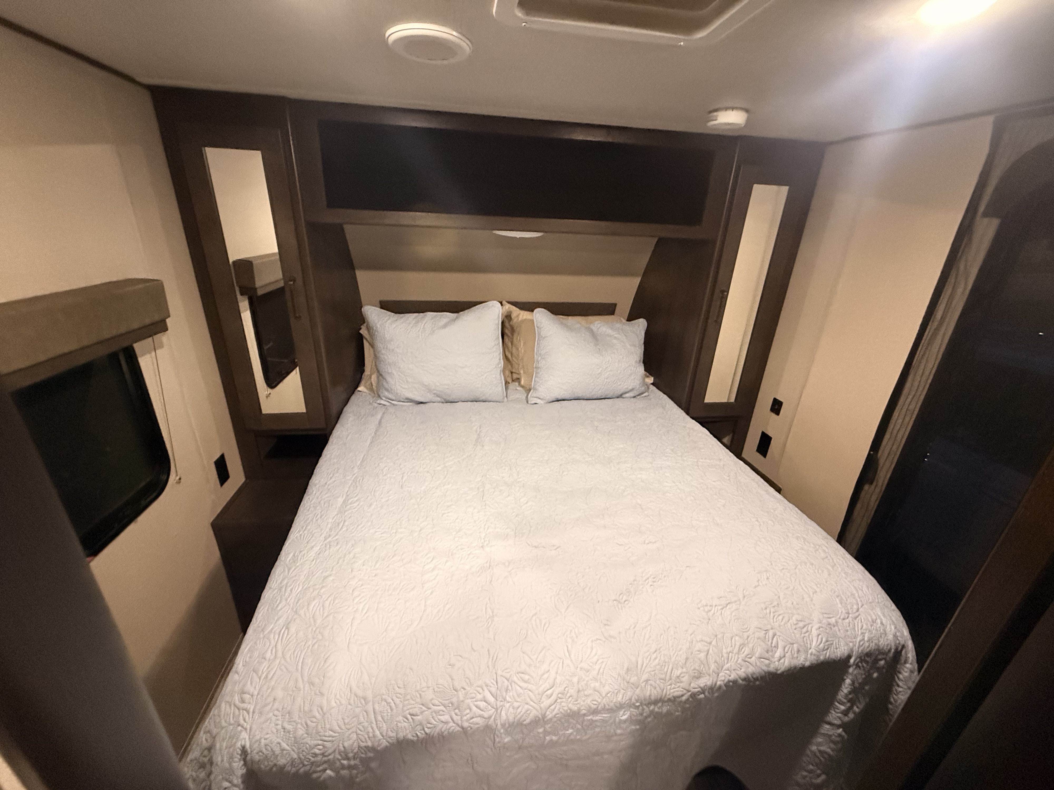 Cozy RV rental bedroom featuring a comfortable bed with pillows, overhead storage cabinets, and a side window.. Grand Design Transcend Xplor 2021