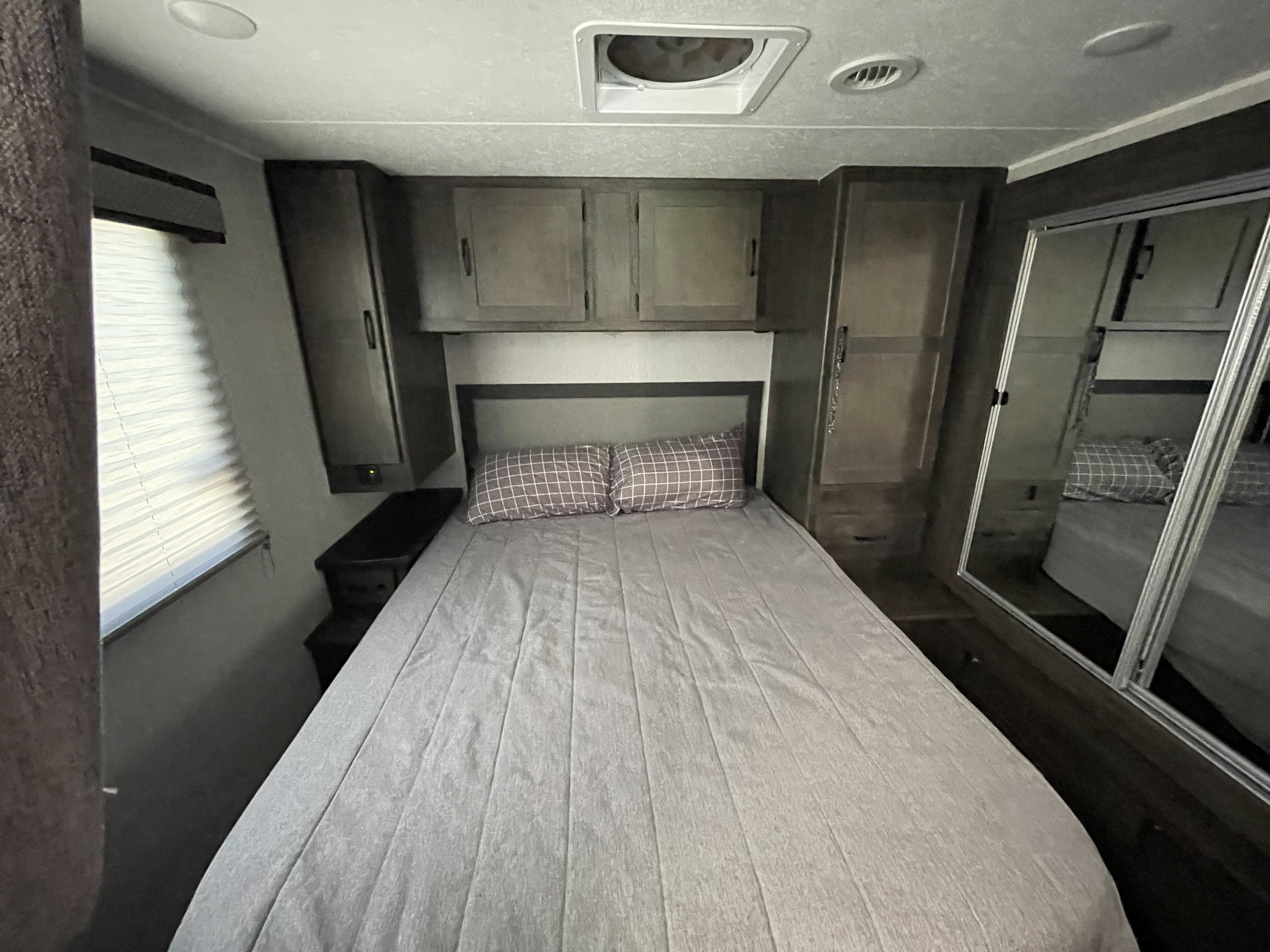 This RV rental bedroom features a comfy bed with checkered pillows, overhead cabinets, side storage, and a mirrored closet.. Forest River Coachmen Leprechaun 2023