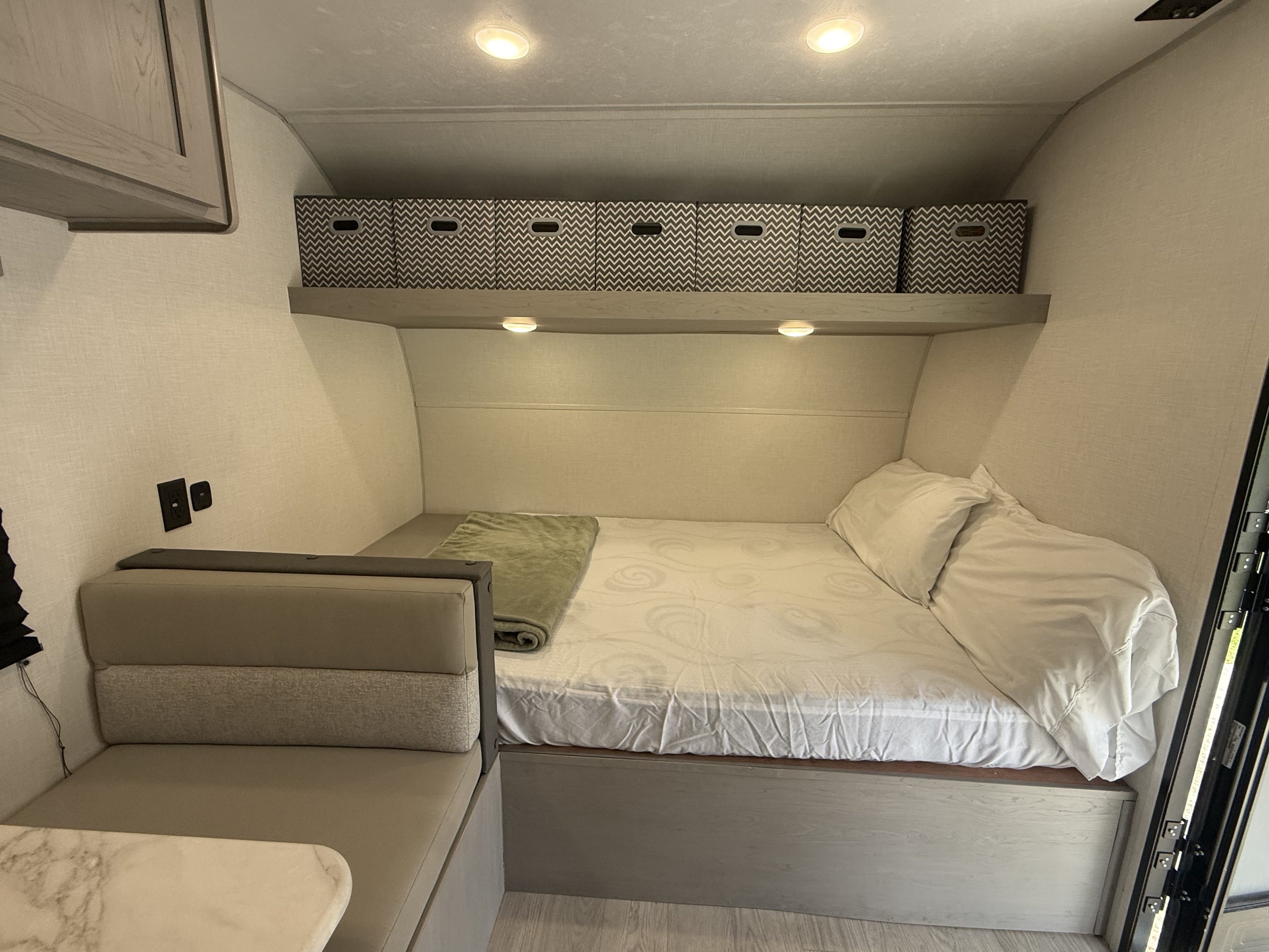 This RV rental interior shows a compact bedroom with a white bed, overhead chevron storage bins, and a dinette.. Coachman Clipper 2026