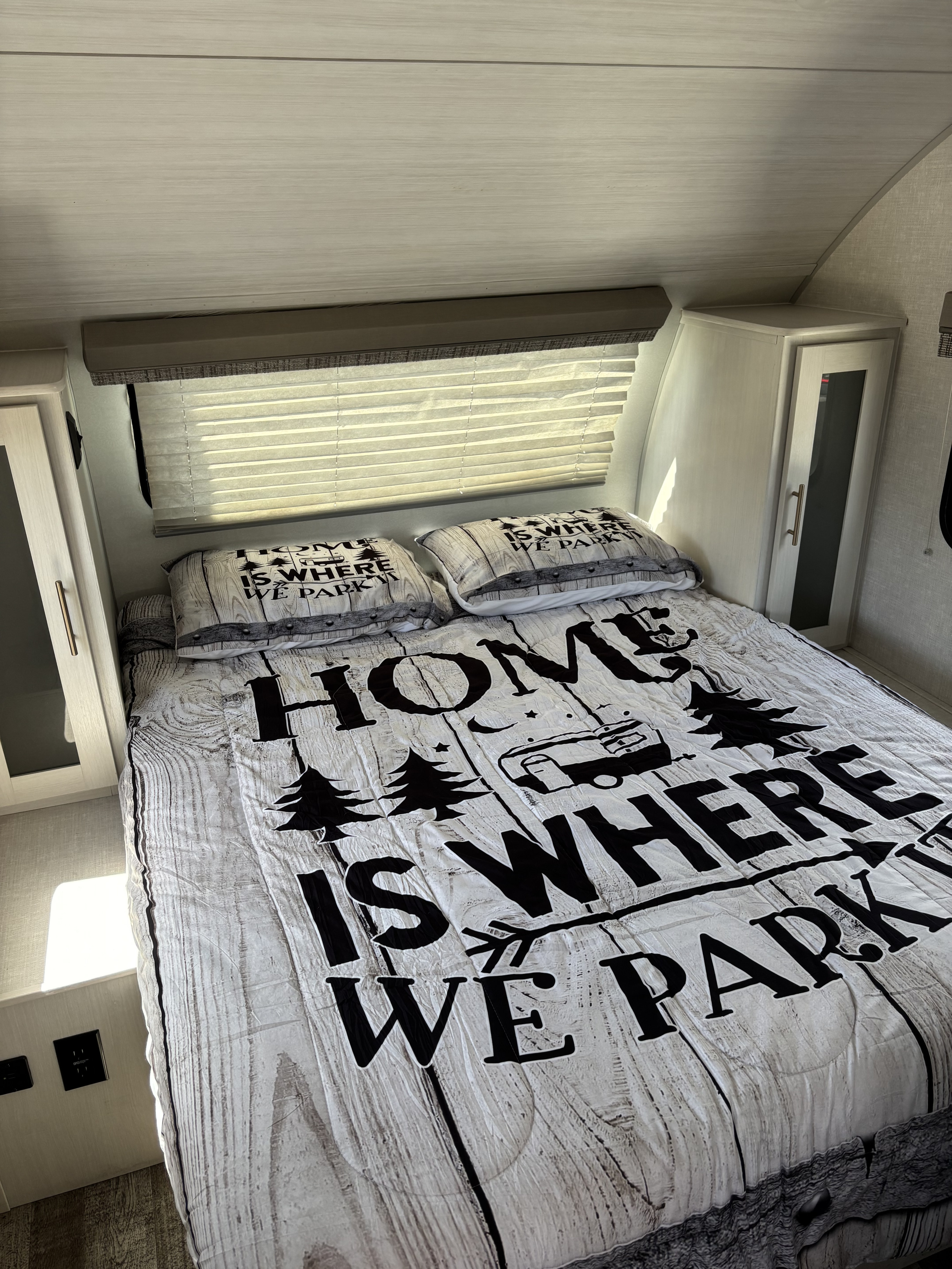 RV rental bedroom with a "Home is where we park it" comforter, side cabinets, and window with blinds.. Forest River 195 RPod 2021