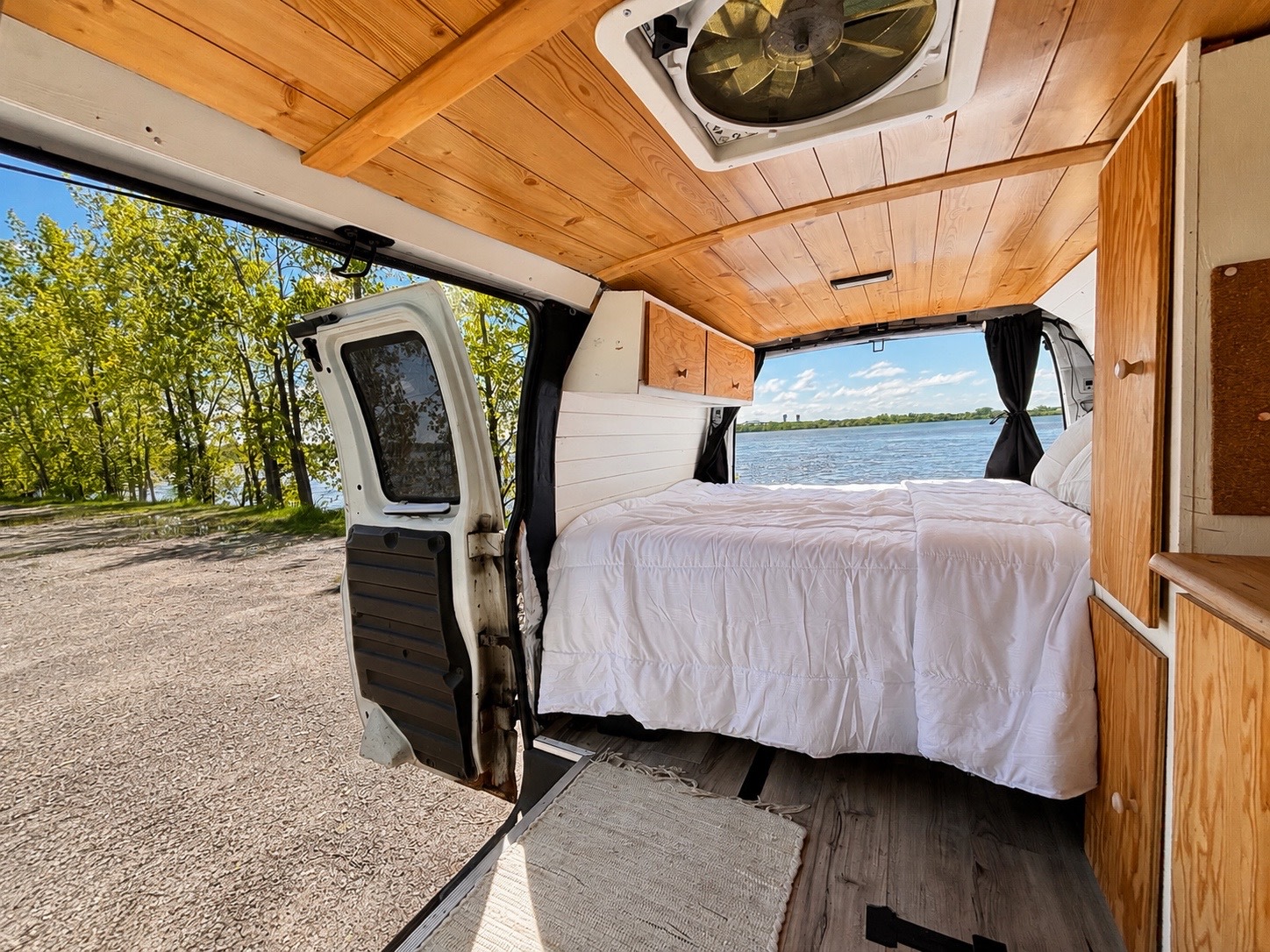Inside an rv rental van, a cozy bed with white duvet, wooden ceiling, and stunning lake view from open rear doors.. Chevrolet Express 2500 2009