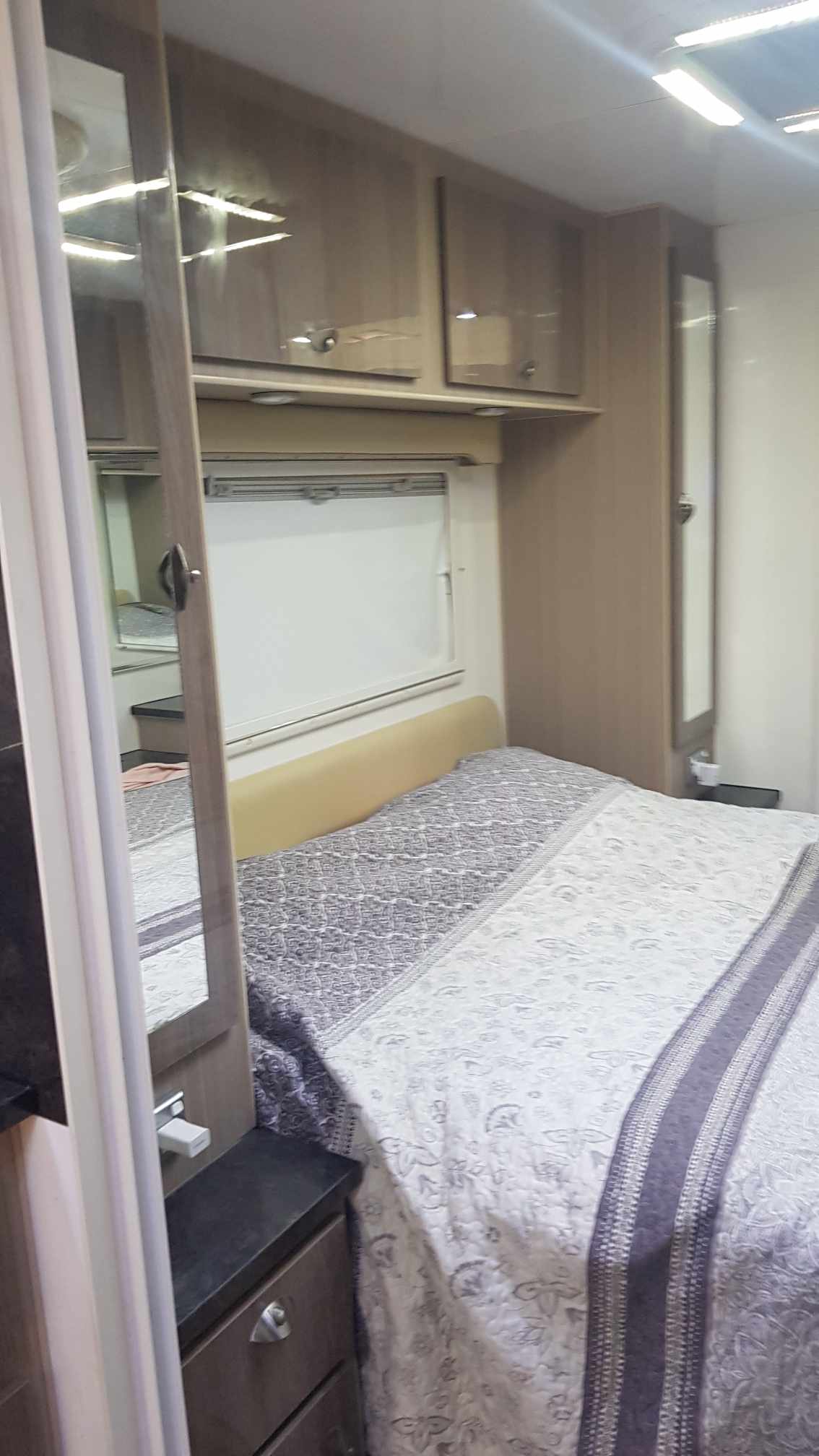 Cozy RV rental bedroom with a patterned bed, wood-toned overhead cabinets, a window, and mirrors. Compact and well-equipped for travel.. Sonic Sonic Trailer 2016