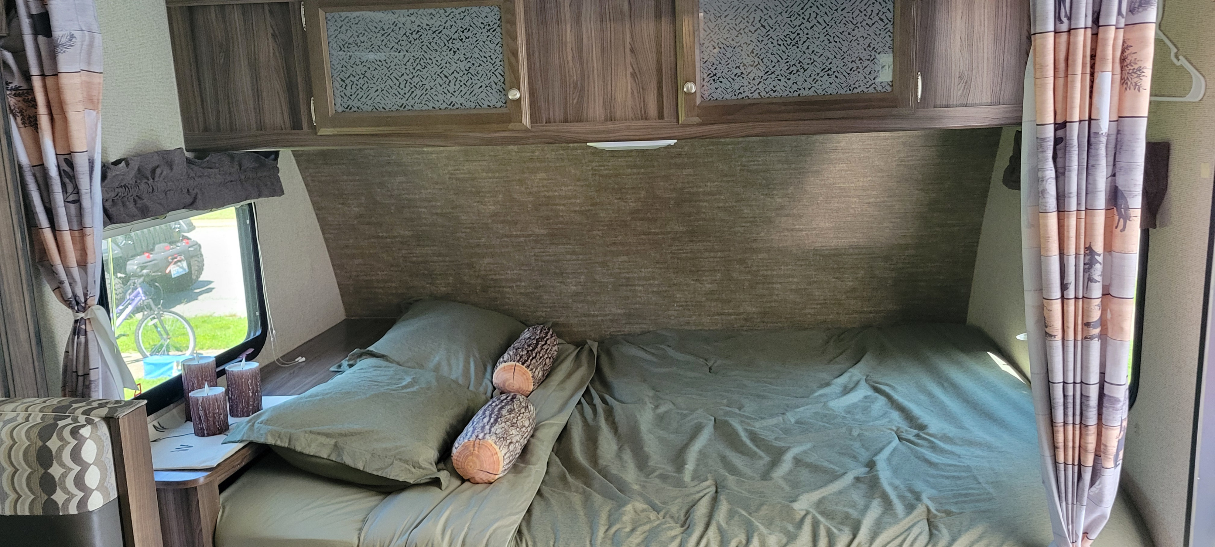 Inside an RV rental: a bed with green sheets, log pillows, overhead cabinets, and a window looking out at an ATV.. Coachmen Apex Nano 2017