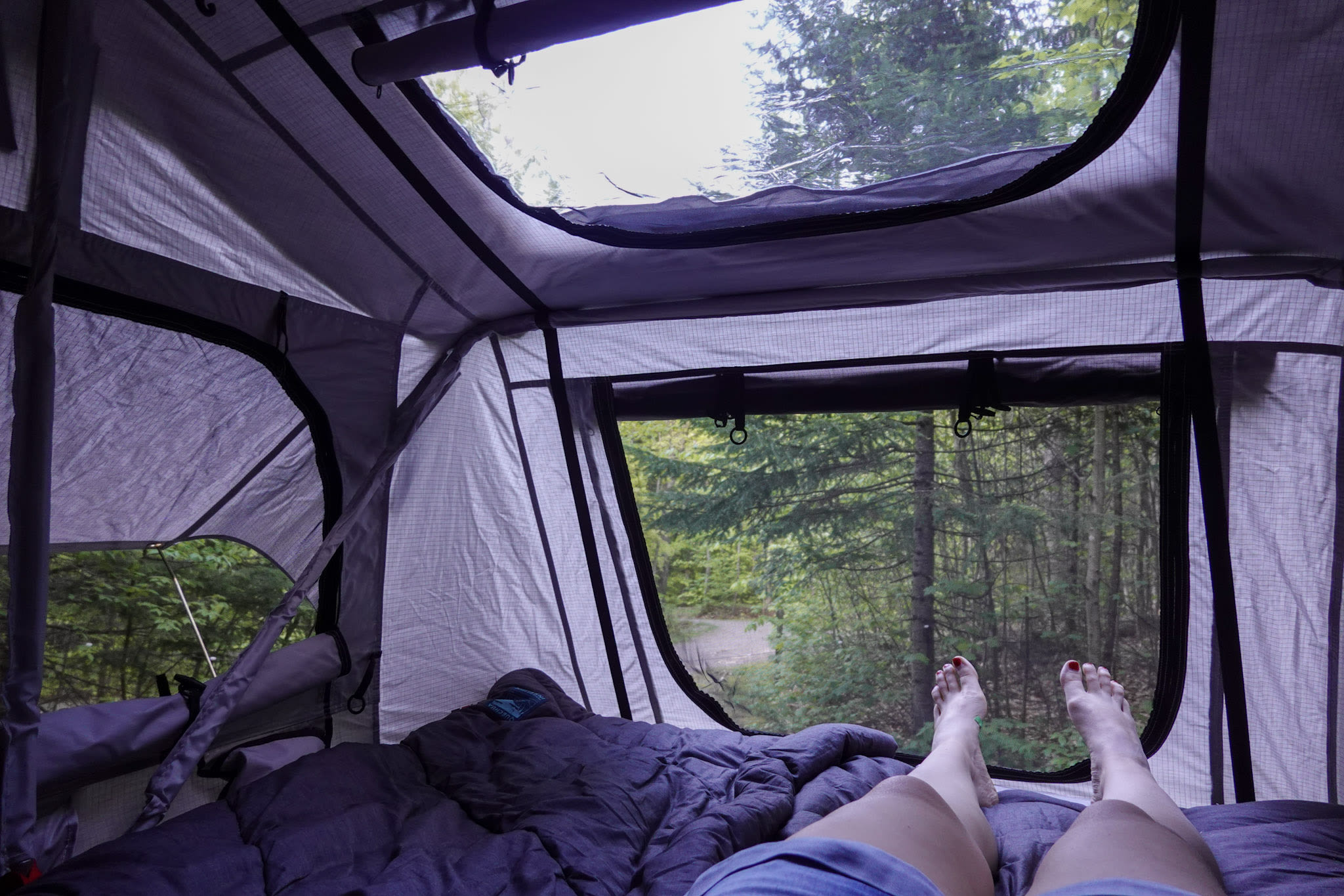 Relaxing in a rooftop tent, bare feet with red nail polish peeking out, enjoying lush forest views. Perfect for an RV rental trip.. Mazda Campervan 2008