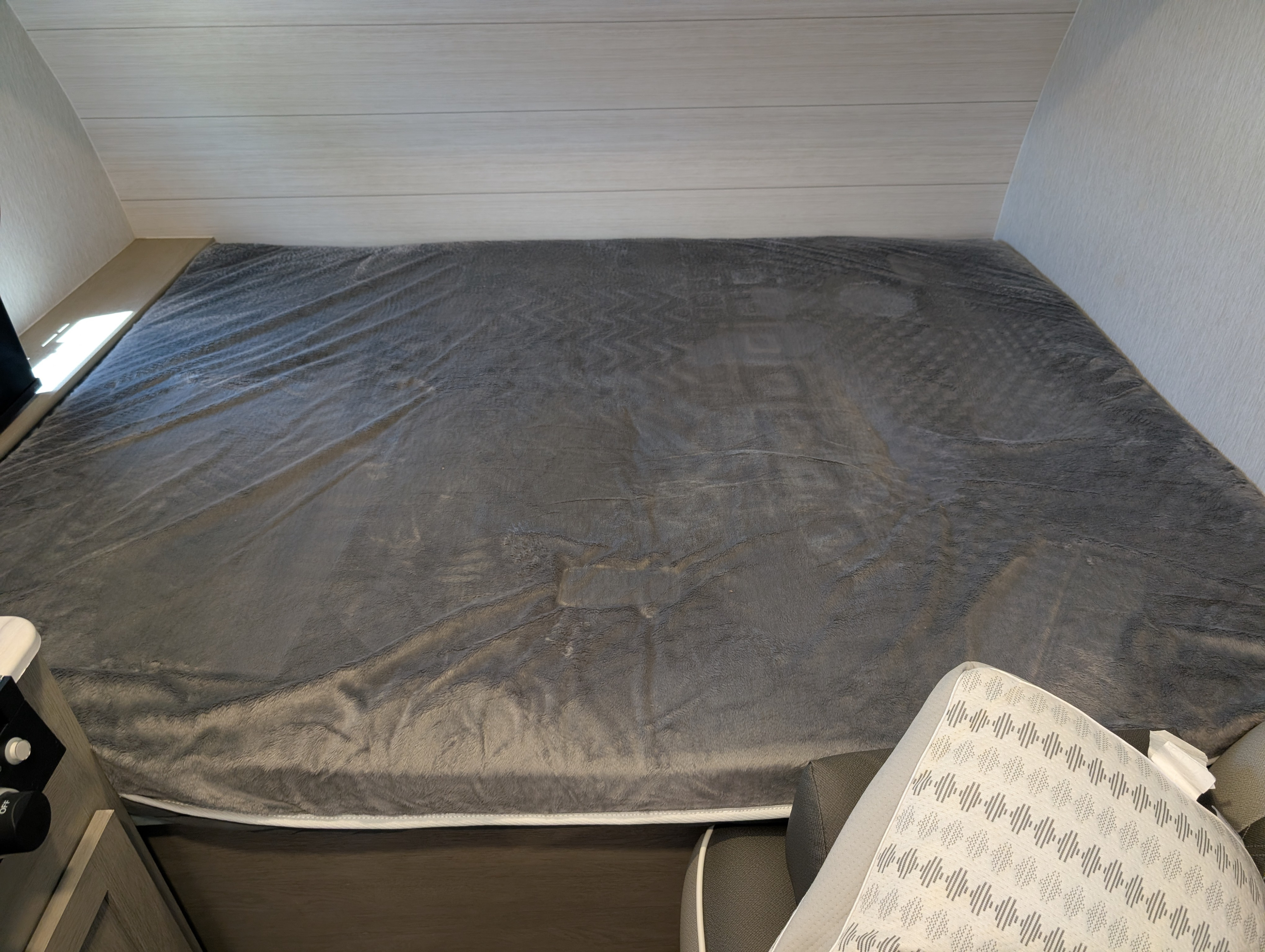 An RV rental's spacious bed with a plush grey blanket, featuring light paneled walls and a cozy, modern camper interior.. Forest River Wildwood 2024