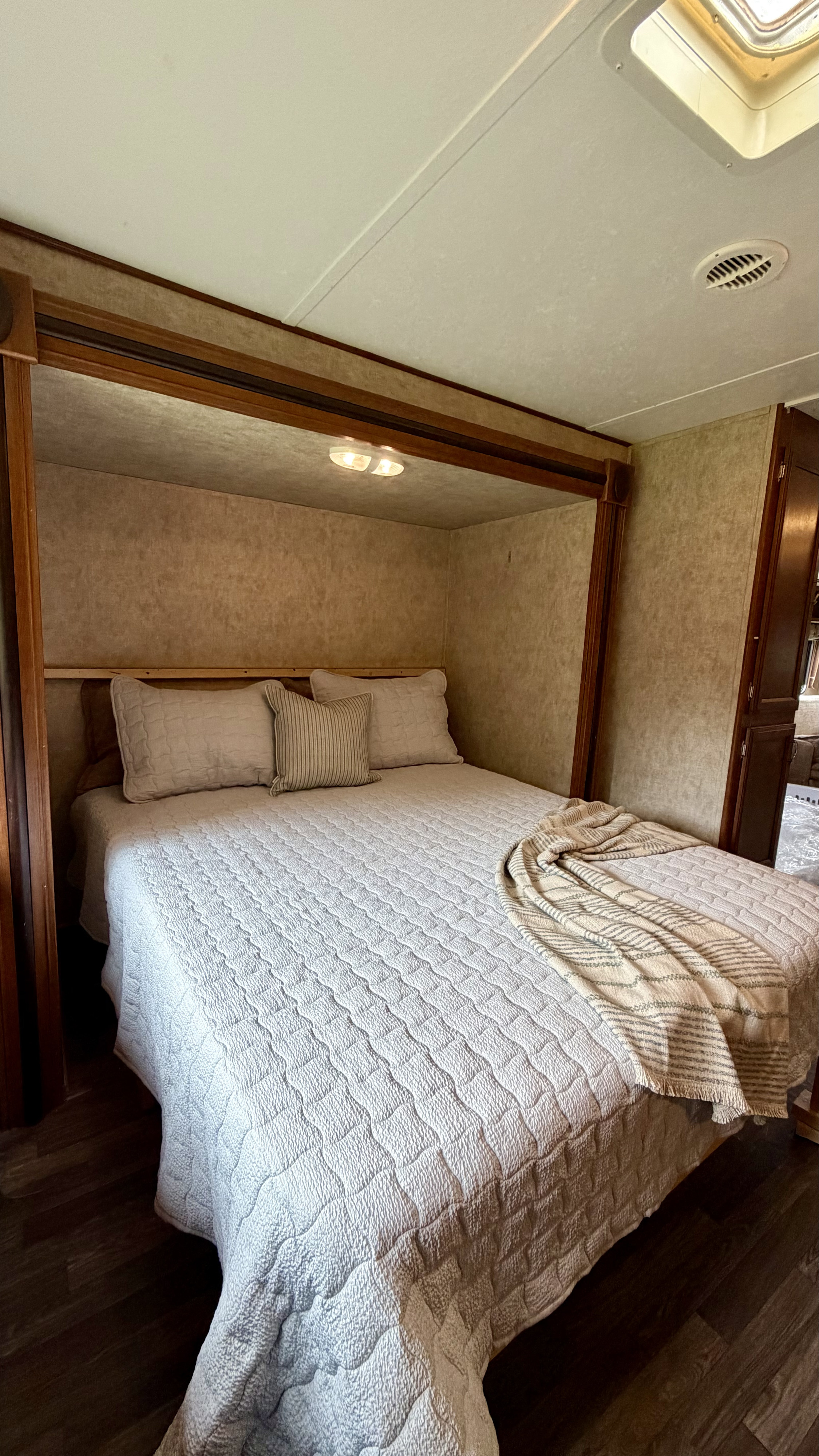 A comfortable RV rental bedroom showing a bed with light-colored quilted bedding, pillows, wood trim, and a ceiling vent.. Avenger Avenger Trailer 2015