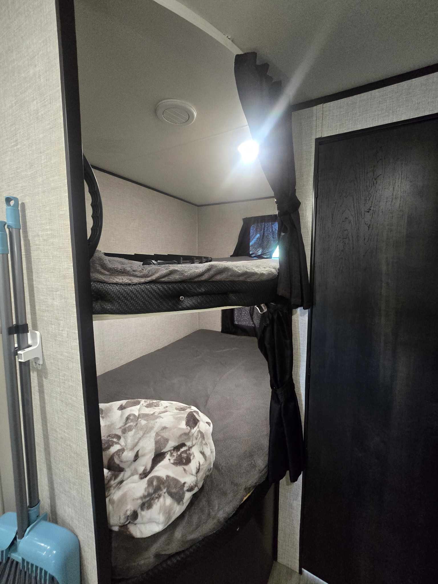 Inside an RV rental: two bunk beds with privacy curtains and a light. A broom set is stored nearby for convenience.. Jayco Jay Flight SLX Rocky Mountain Edition 2022