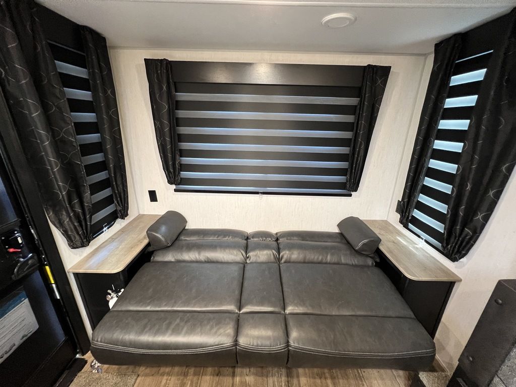 RV rental interior features a dark gray convertible couch/bed, flanked by windows with horizontal blinds and black curtains.. Forest River Cherokee Grey Wolf 2022