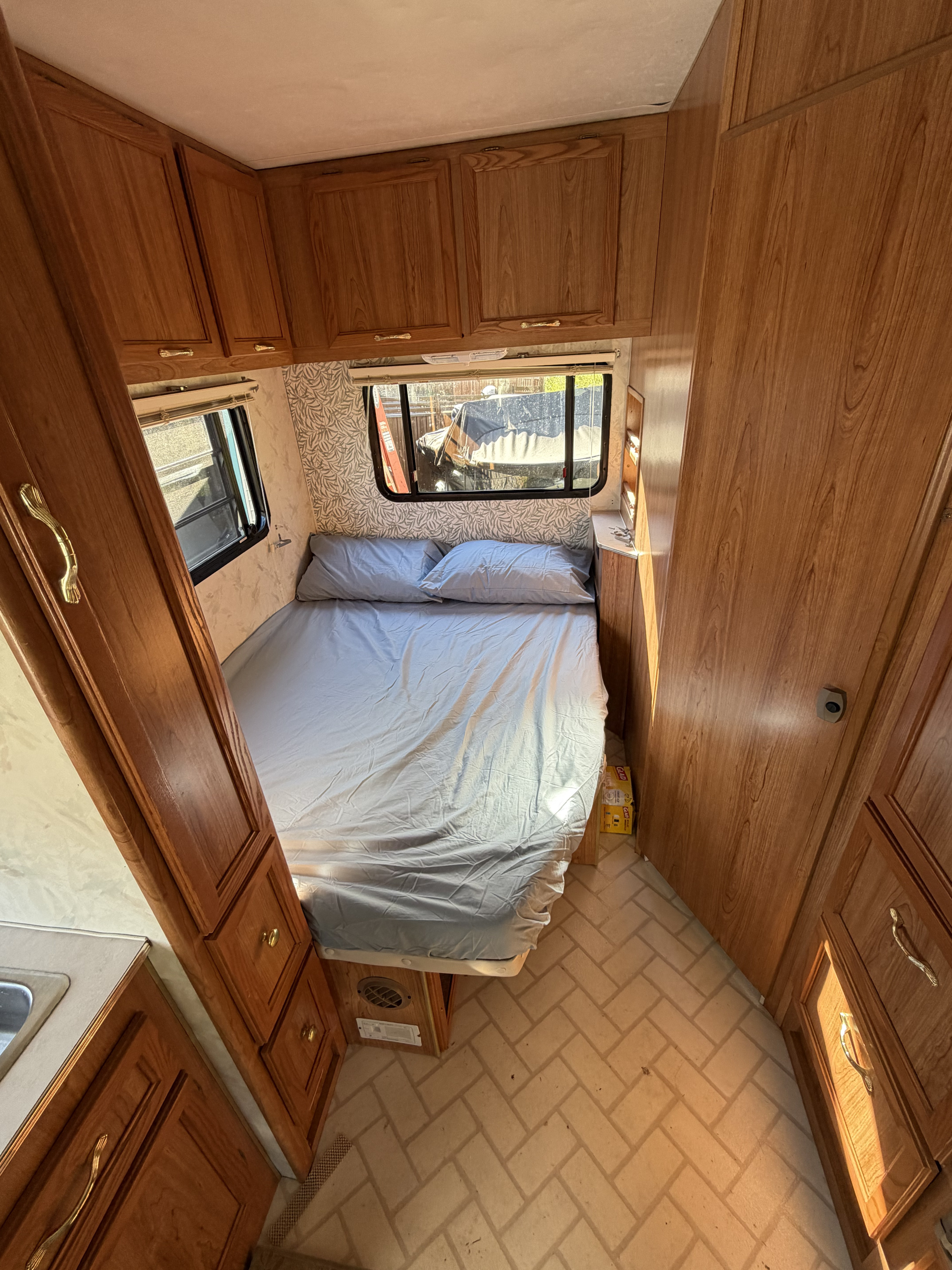 Cozy RV rental bedroom featuring a bed with light blue bedding, wooden cabinets, patterned walls, and sunlit windows.. Ford Rv E-350/Catalina 1997