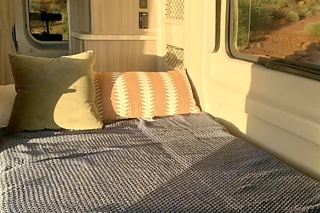 Inside a cozy RV rental: a bed with olive and patterned pillows, blue waffle blanket, window view of a desert landscape.. Thor Scope 2023