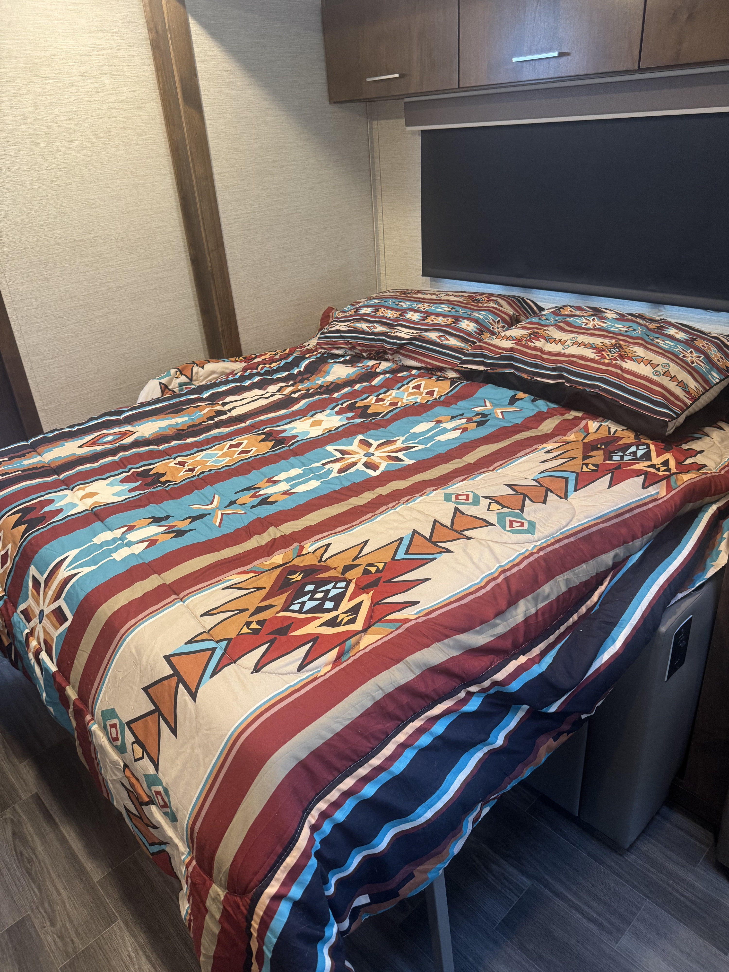 Cozy RV rental bedroom featuring a colorful Southwestern bed, overhead cabinets, and wood-look floor.. Wayfarer Tiffin 2021