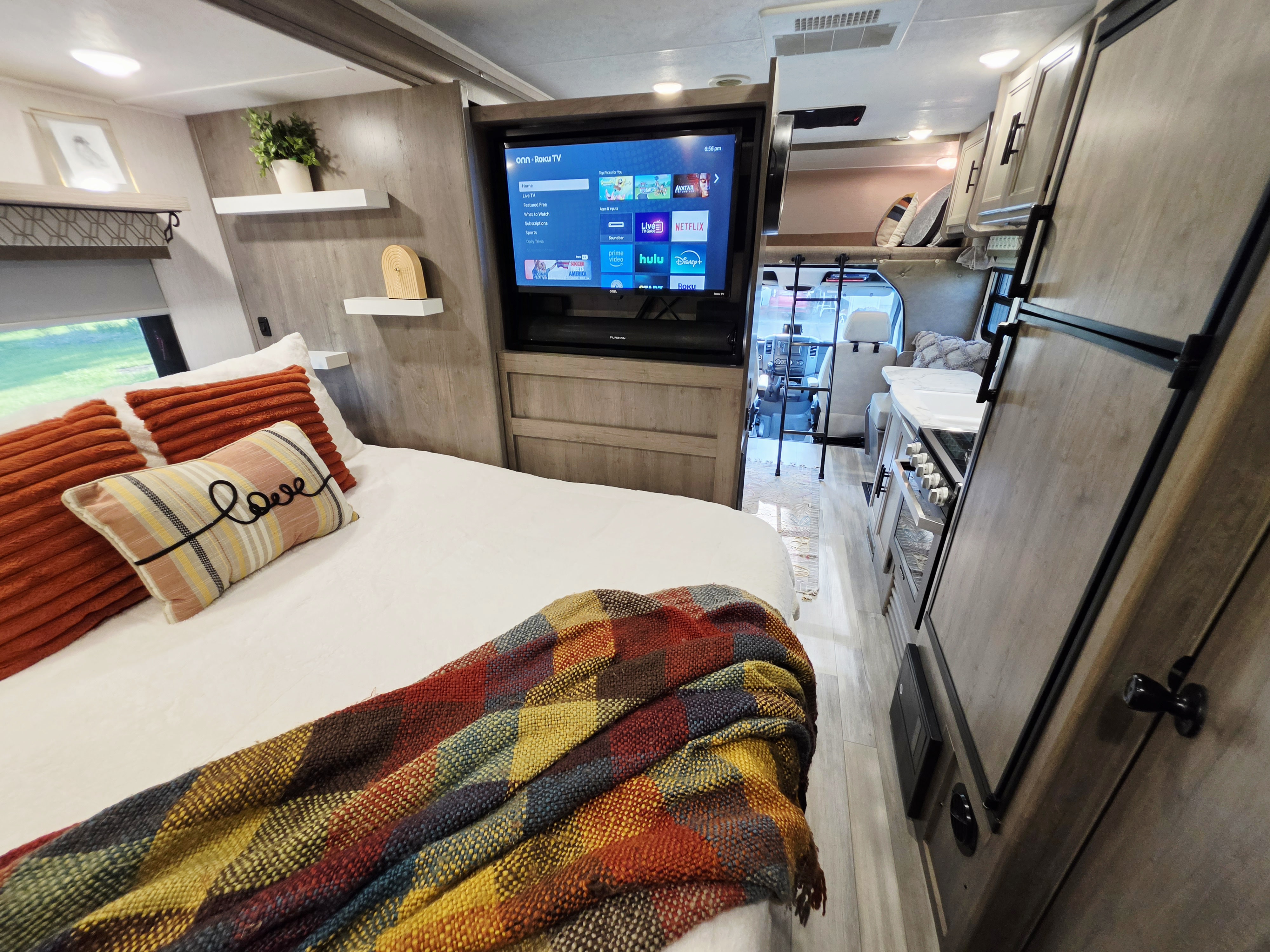 Cozy rv rental bedroom features a smart Roku TV, colorful bedding, and a glimpse into the kitchen and driver's cab.. Coachmen Prism 2018