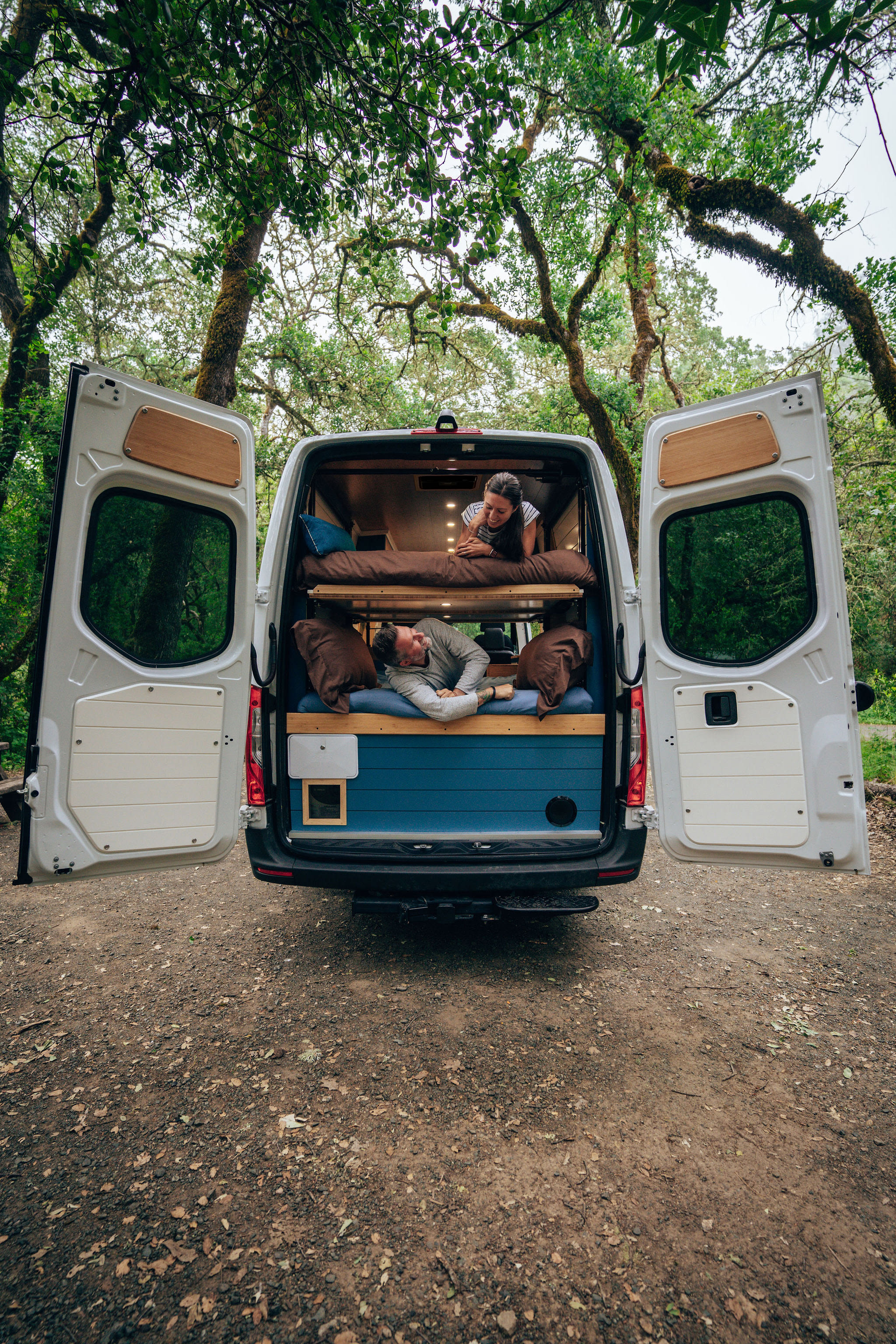 A couple relaxes in their RV rental camper van with bunk beds, rear doors open to a lush forest. Perfect for adventure.. Mercedes-Benz Sprinter 2024