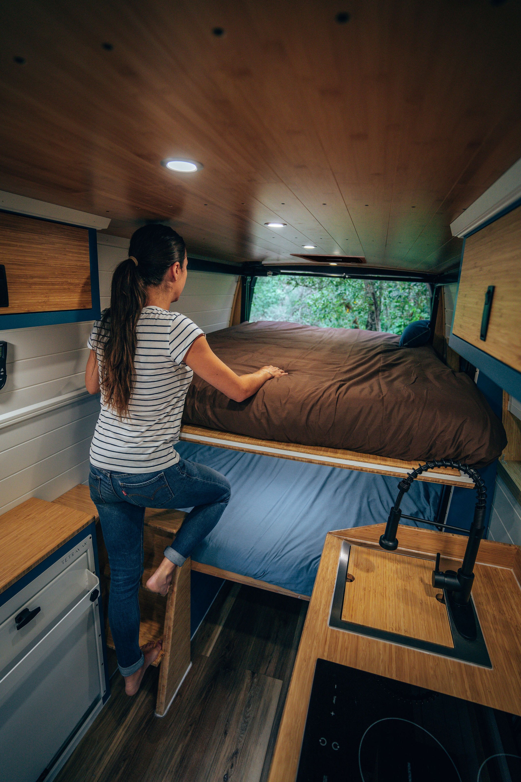 RV rental interior: Woman stepping into an elevated bed in a cozy camper van, featuring a kitchen and forested view.. Mercedes-Benz Sprinter 2024
