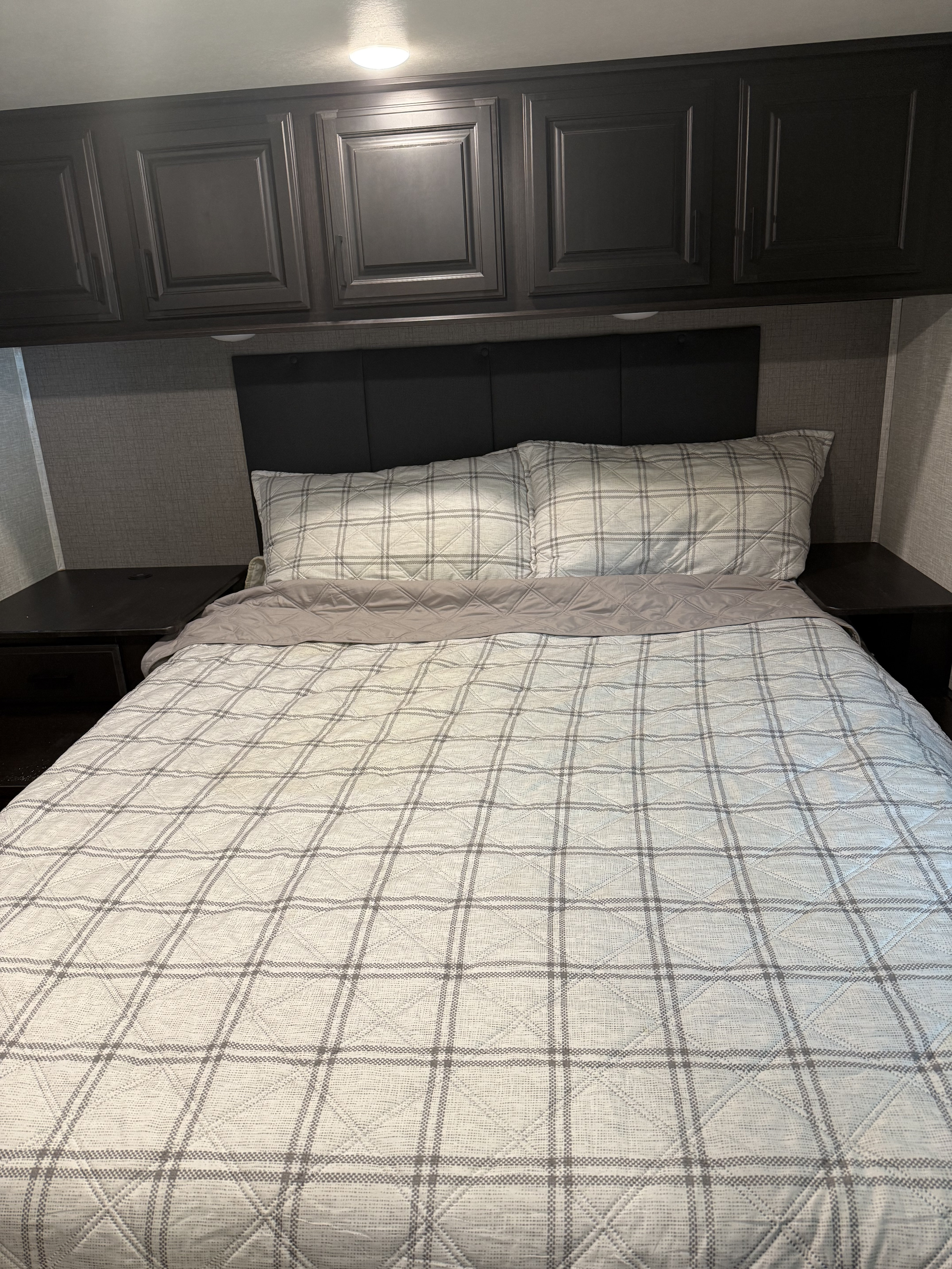 Cozy RV rental bedroom featuring a plaid bed, dark headboard, side tables, and overhead cabinets for storage.. Highland Ridge RV Open Range 2023