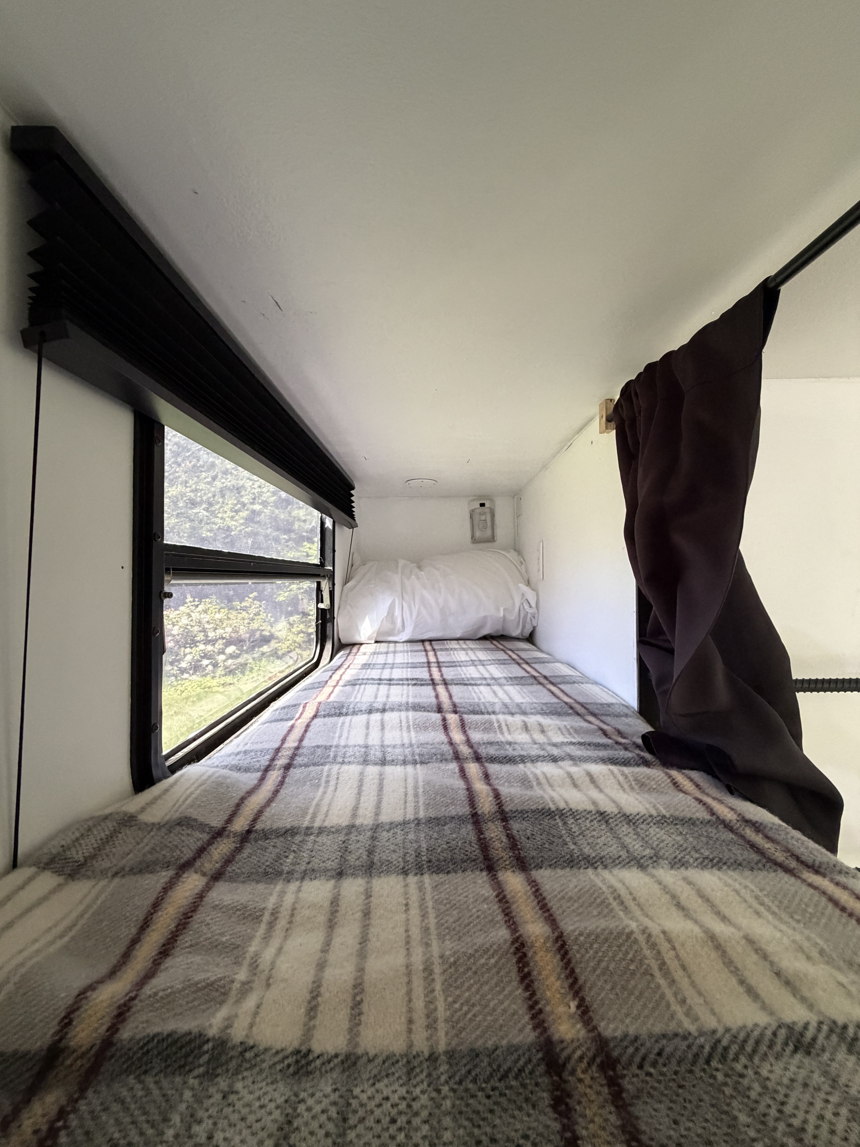 Inside an RV rental: a cozy, narrow sleeping bunk with plaid blanket, pillow, window view, and dark curtain.. Thor Motor Coach Citation 1999