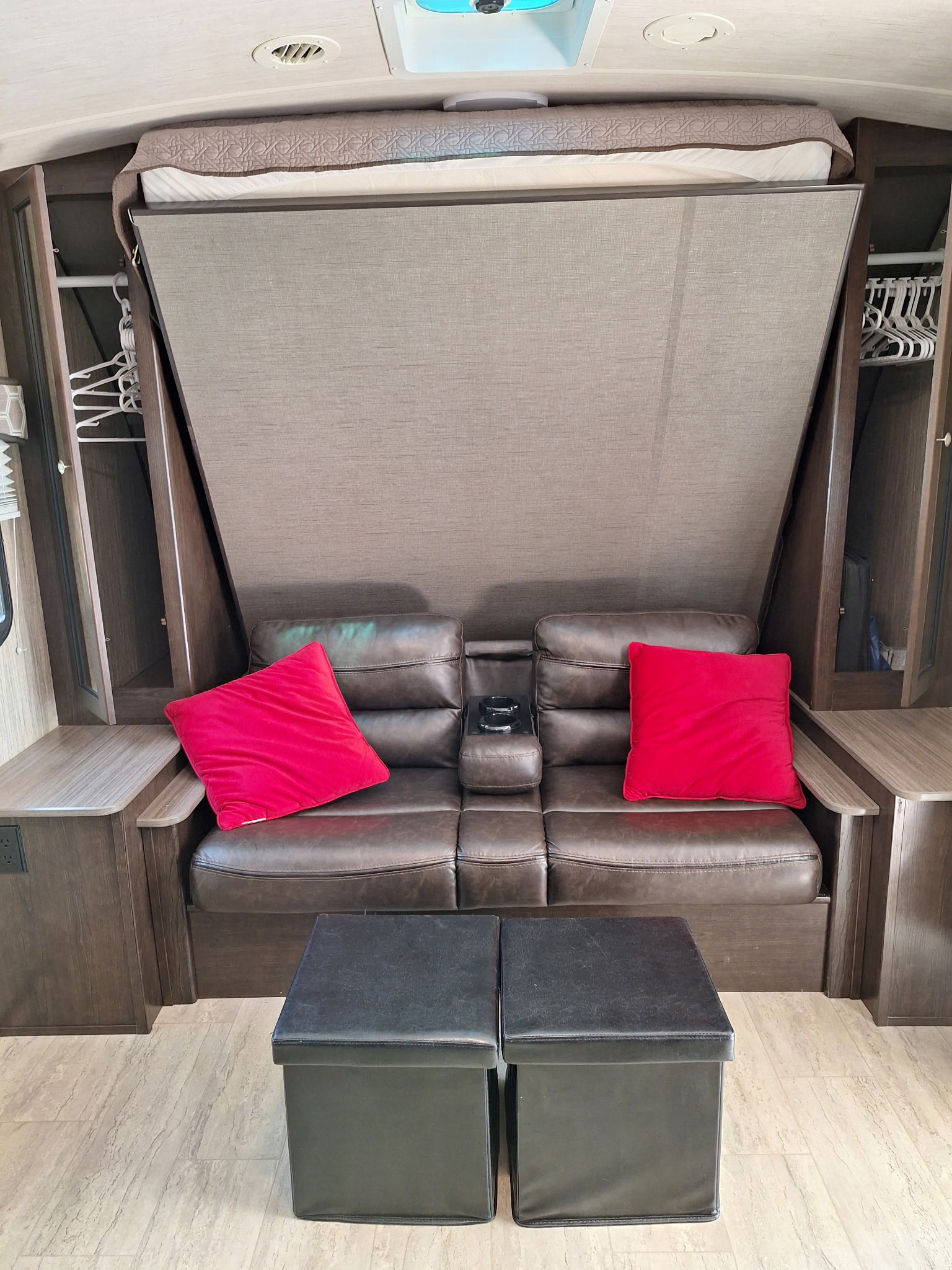 RV rental interior features a folded-up Murphy bed over a brown sofa with red cushions and two black storage ottomans.. Cruiser RV Shadow Cruiser 2018