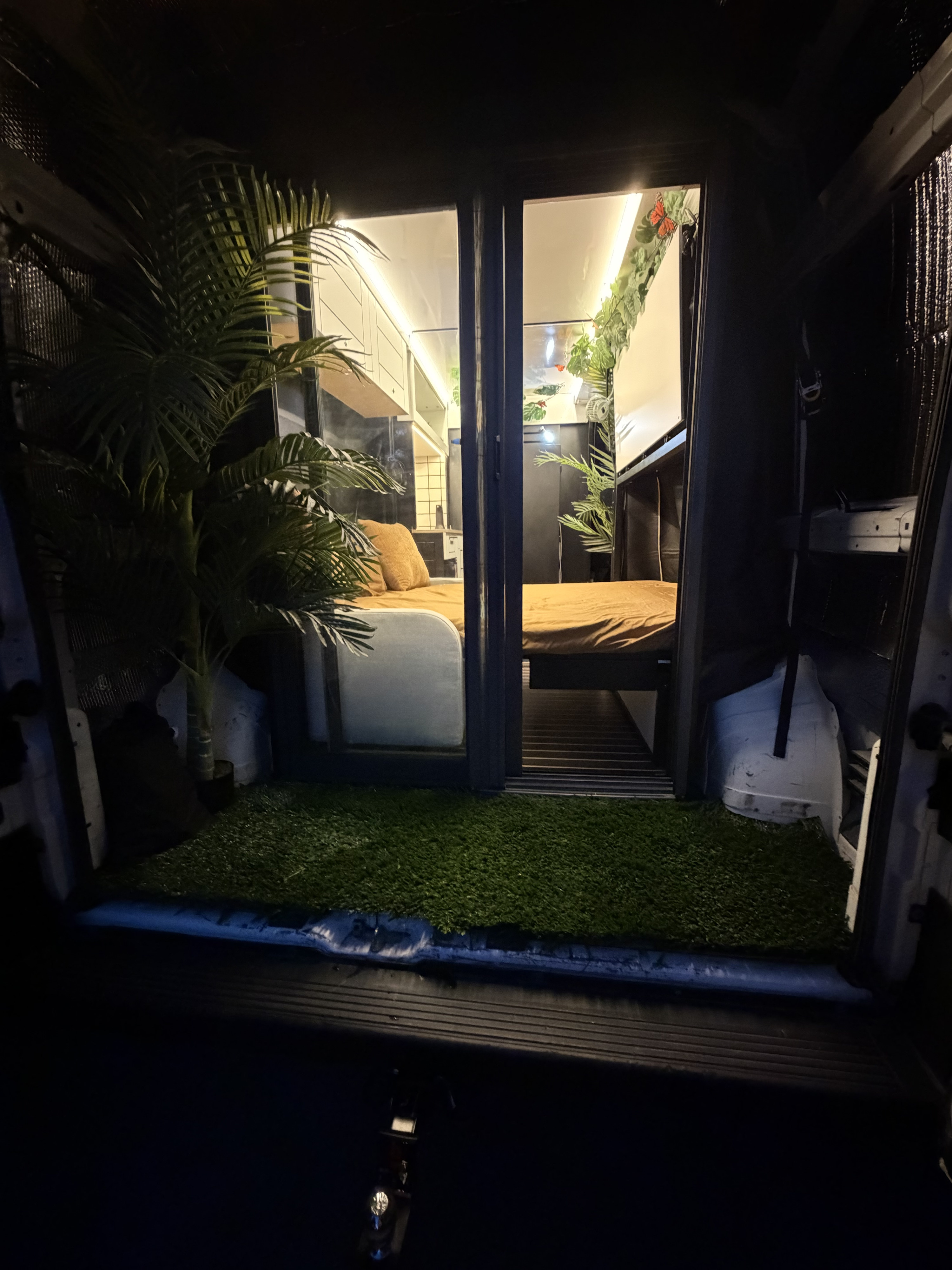 Night view of an RV rental's inviting interior: two beds, plants, strip lighting, and a soft artificial grass entrance.. NA NA 2021