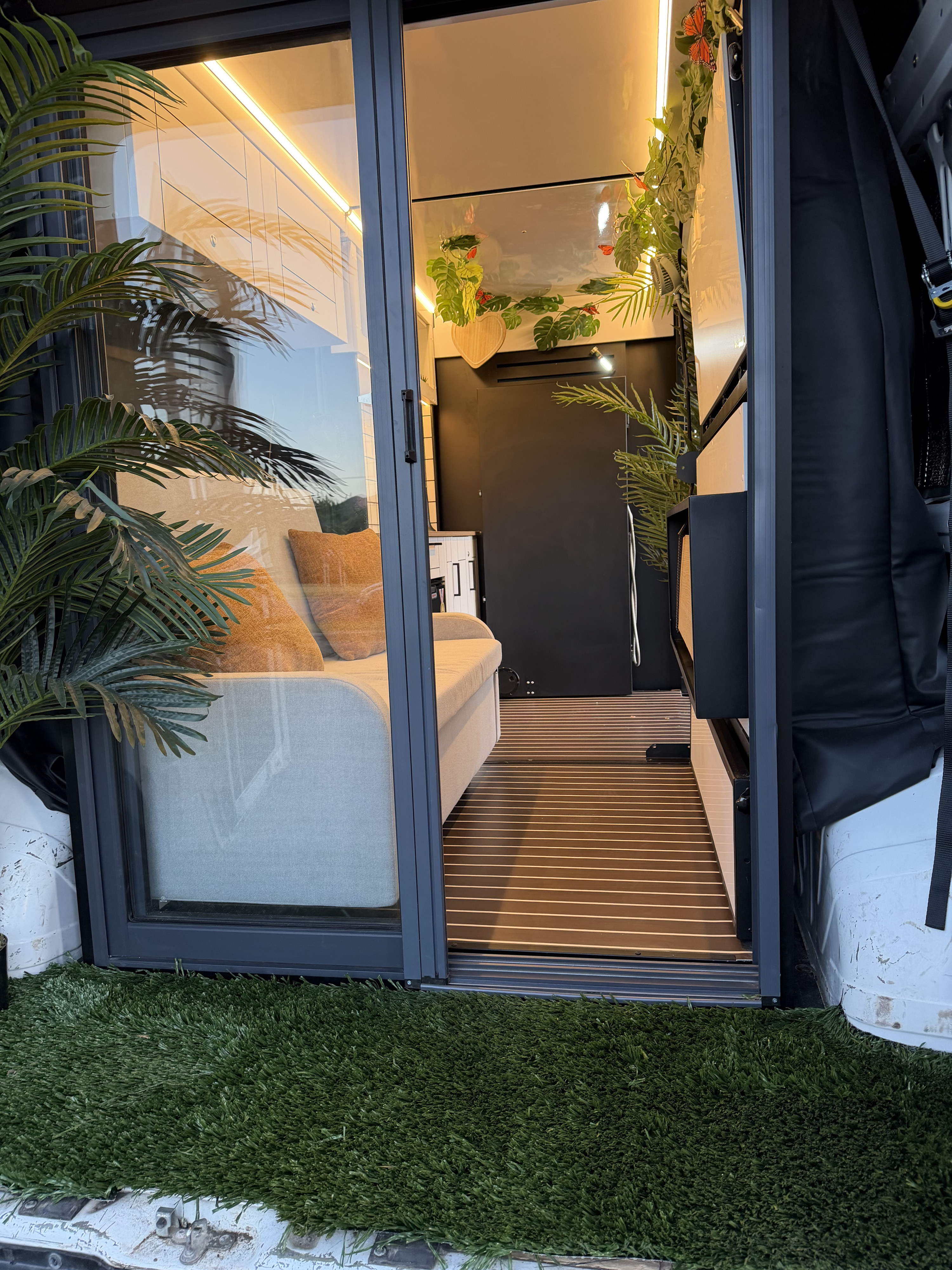 View of a modern rv rental's cozy camper van interior through a glass door, featuring warm lighting, green plants, and a comfortable couch.. NA NA 2021