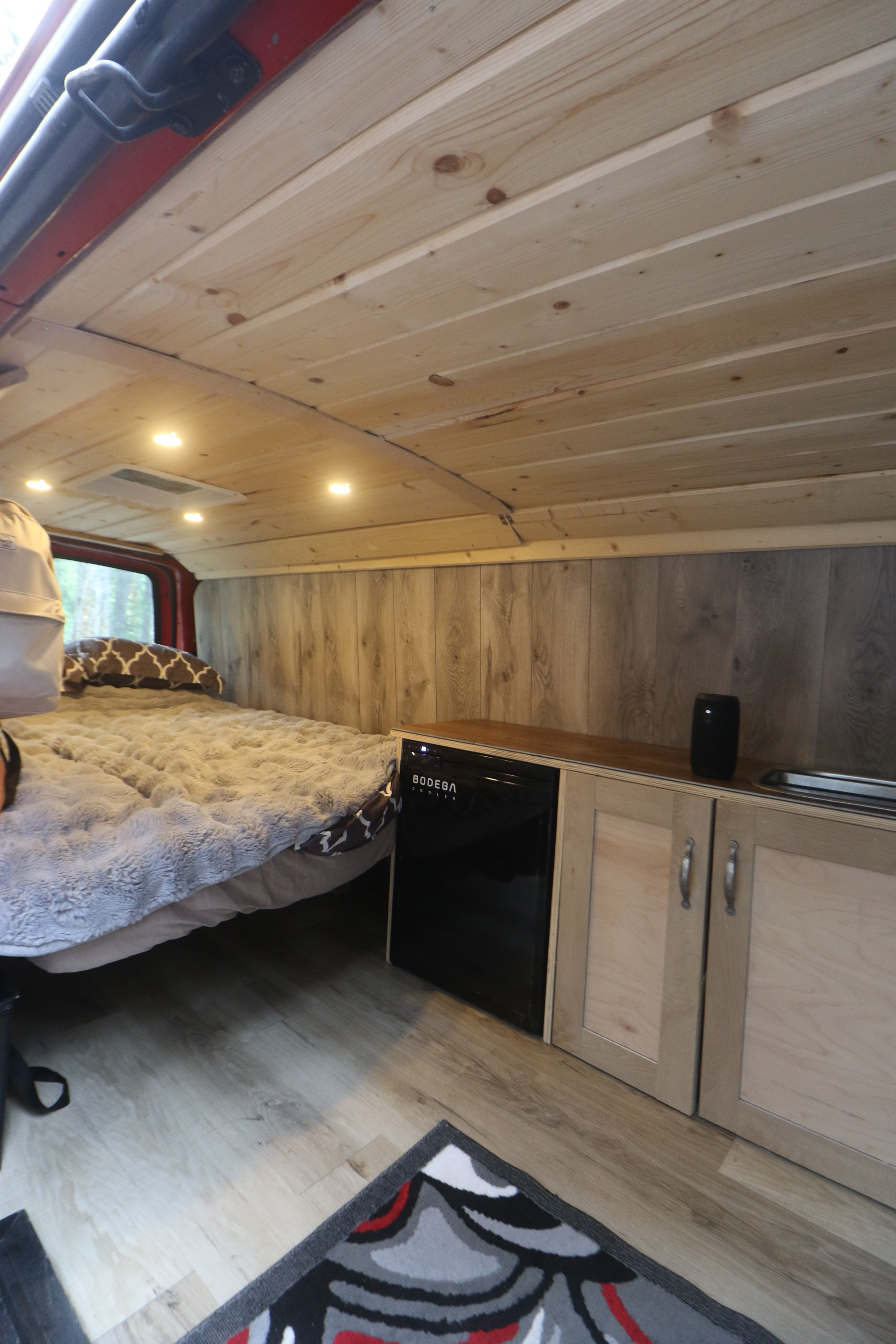 Cozy RV rental interior: wooden ceiling, comfy bed, kitchenette with mini-fridge & sink, plus wood flooring.. Glacier Campers Vanlife Explorer 2013