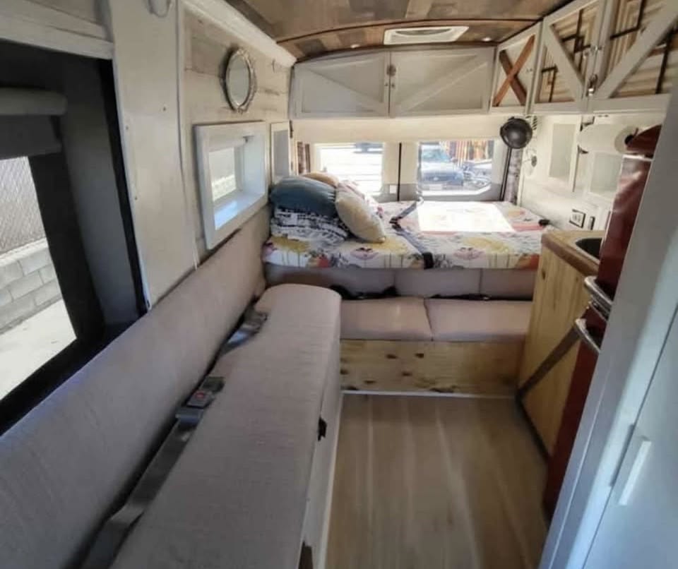 RV rental interior showcasing a cozy bed, long bench seat with seatbelts, wooden floor & ceiling, and white storage cabinets.. Ford T-350 Transit Wagon 2016