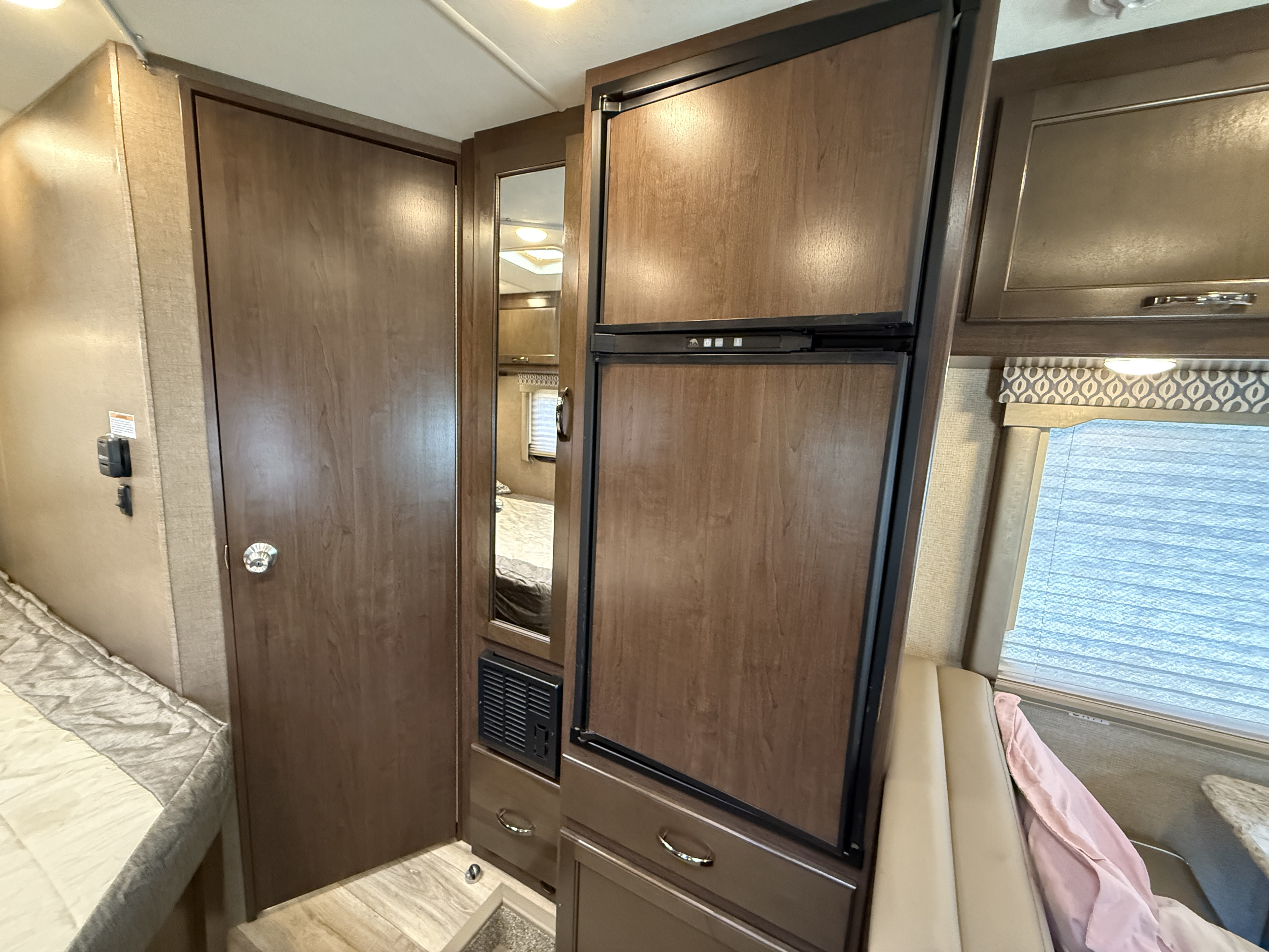 RV rental interior: wood-paneled fridge, door to bedroom/bath, mirrored cabinet, bed, window, and dinette seating.. Thor Freedom Elite 2018