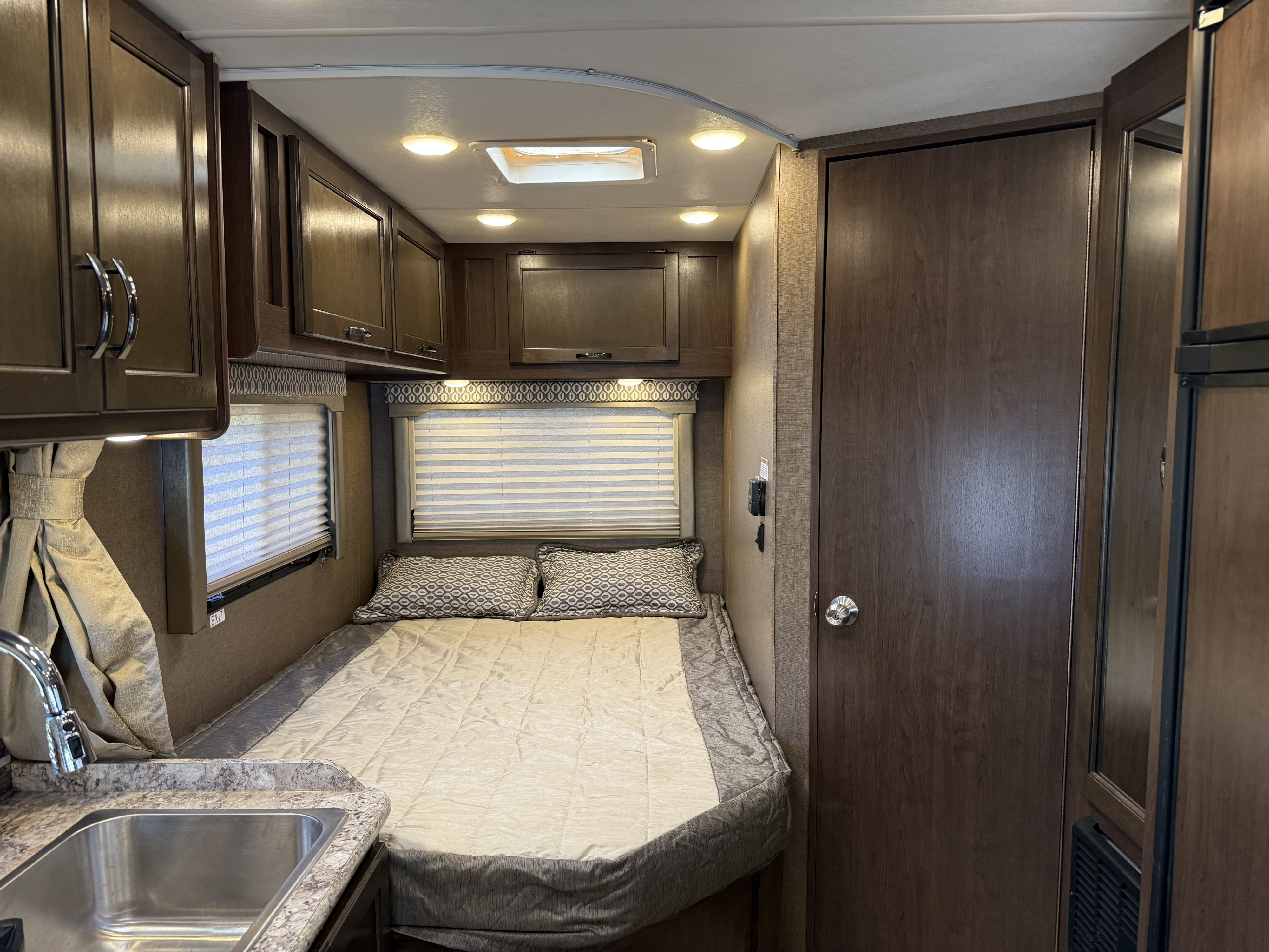 Inside an RV rental, a compact bedroom area with a bed, windows, overhead cabinets, and a sink on the left.. Thor Freedom Elite 2018