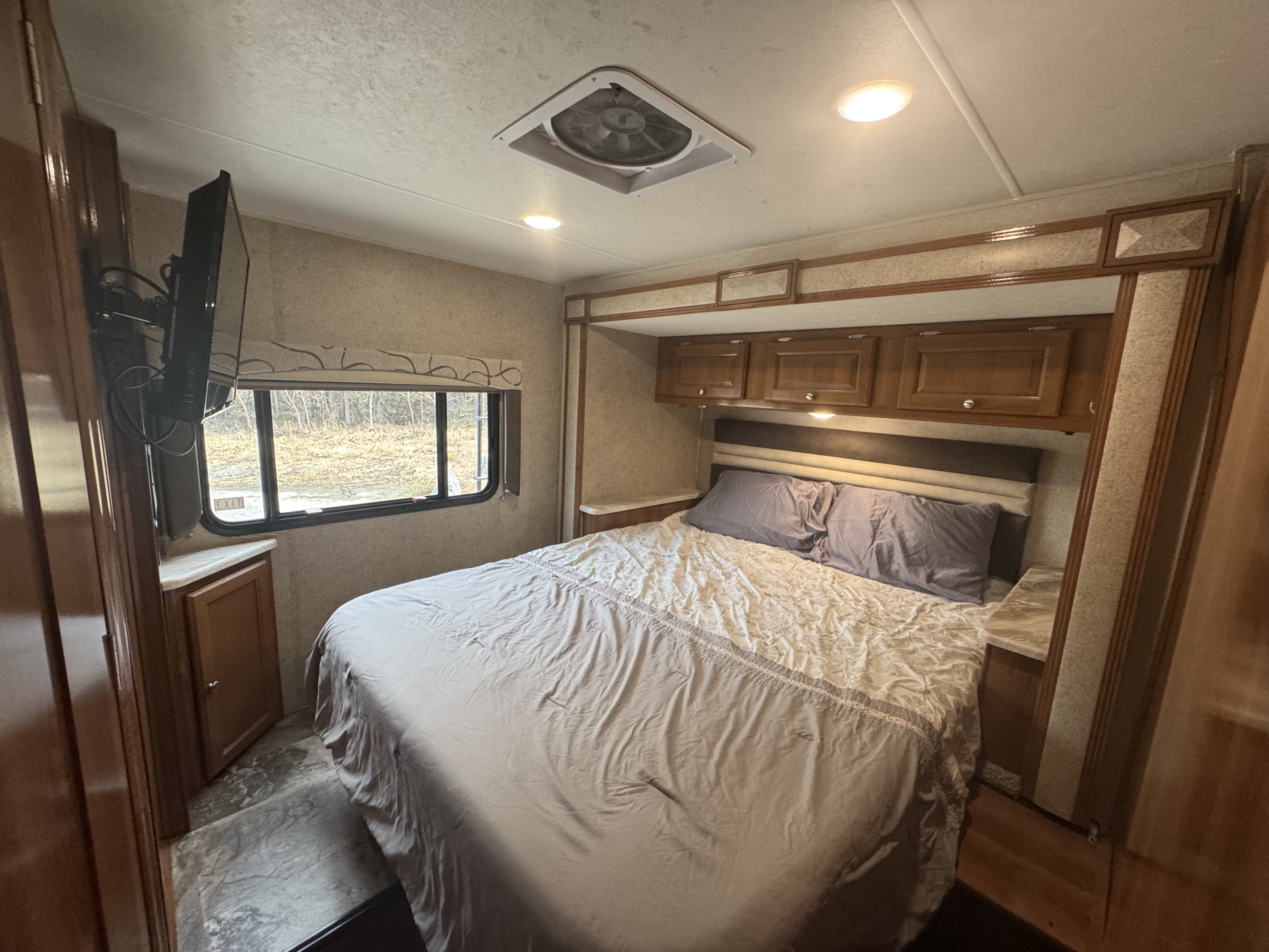RV rental bedroom with queen bed, overhead storage, wall-mounted TV, and a window offering outdoor views for a comfortable stay.. Coachmen Leprechaun 260DS 2019