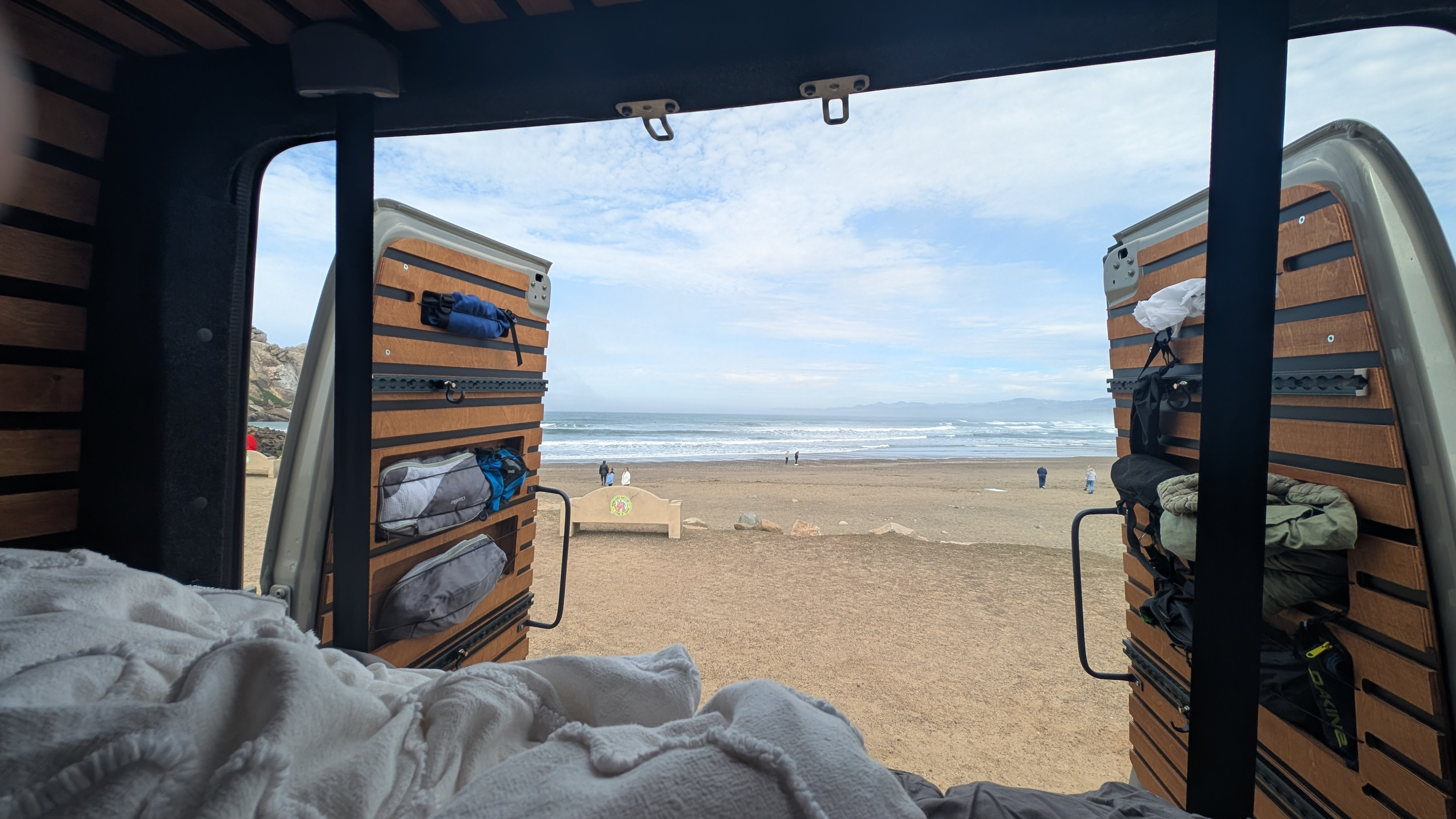 Cozy rv rental view from bed: ocean waves, sandy beach, and distant hills seen through open camper van doors.. Mercedes-Benz Sprinter RV Motorhome Campervan 2023