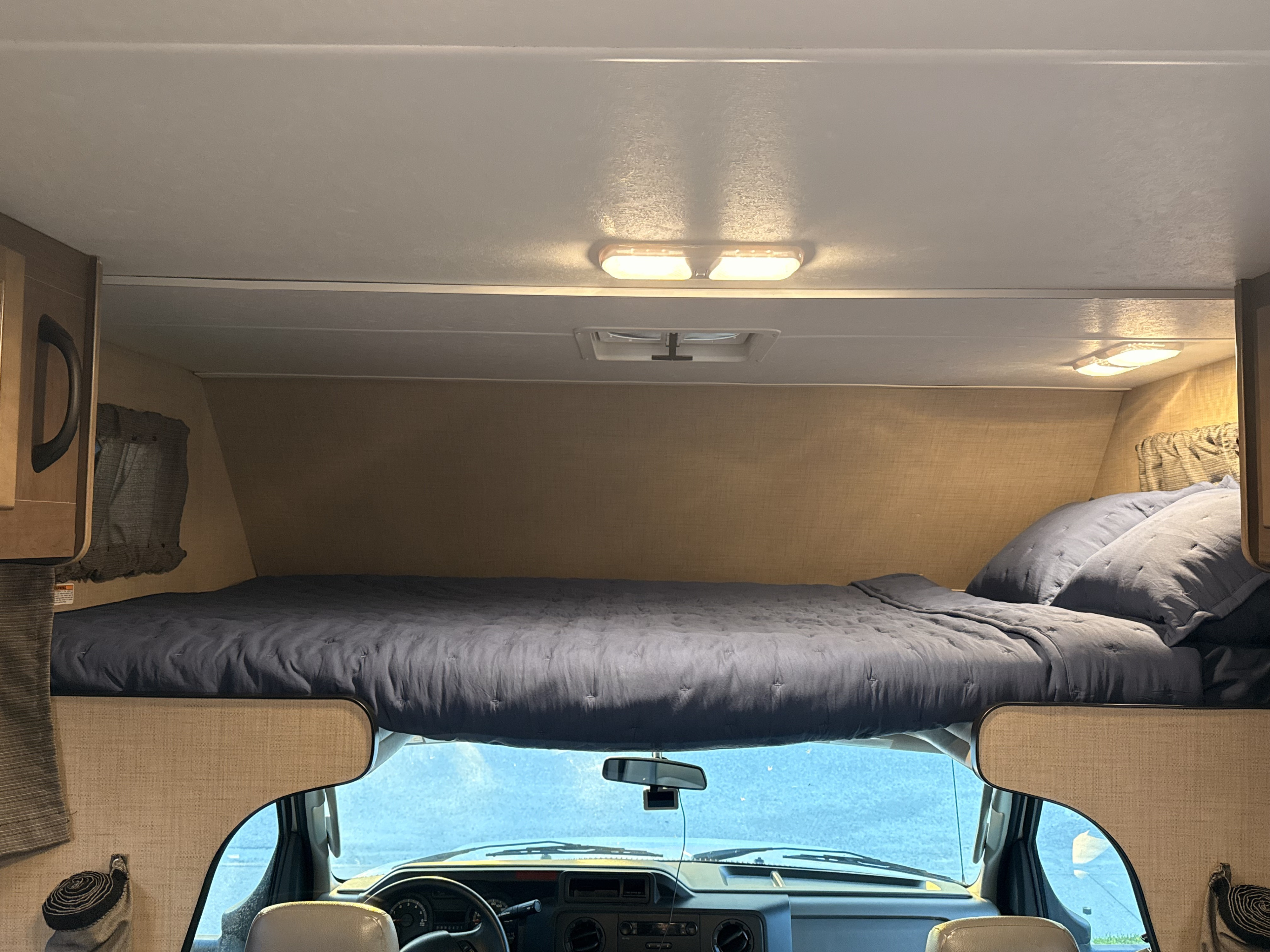 Inside an rv rental: a cozy cab-over bunk bed with blue bedding, overlooking the driver's area and windshield.. Thor Majestic 2020