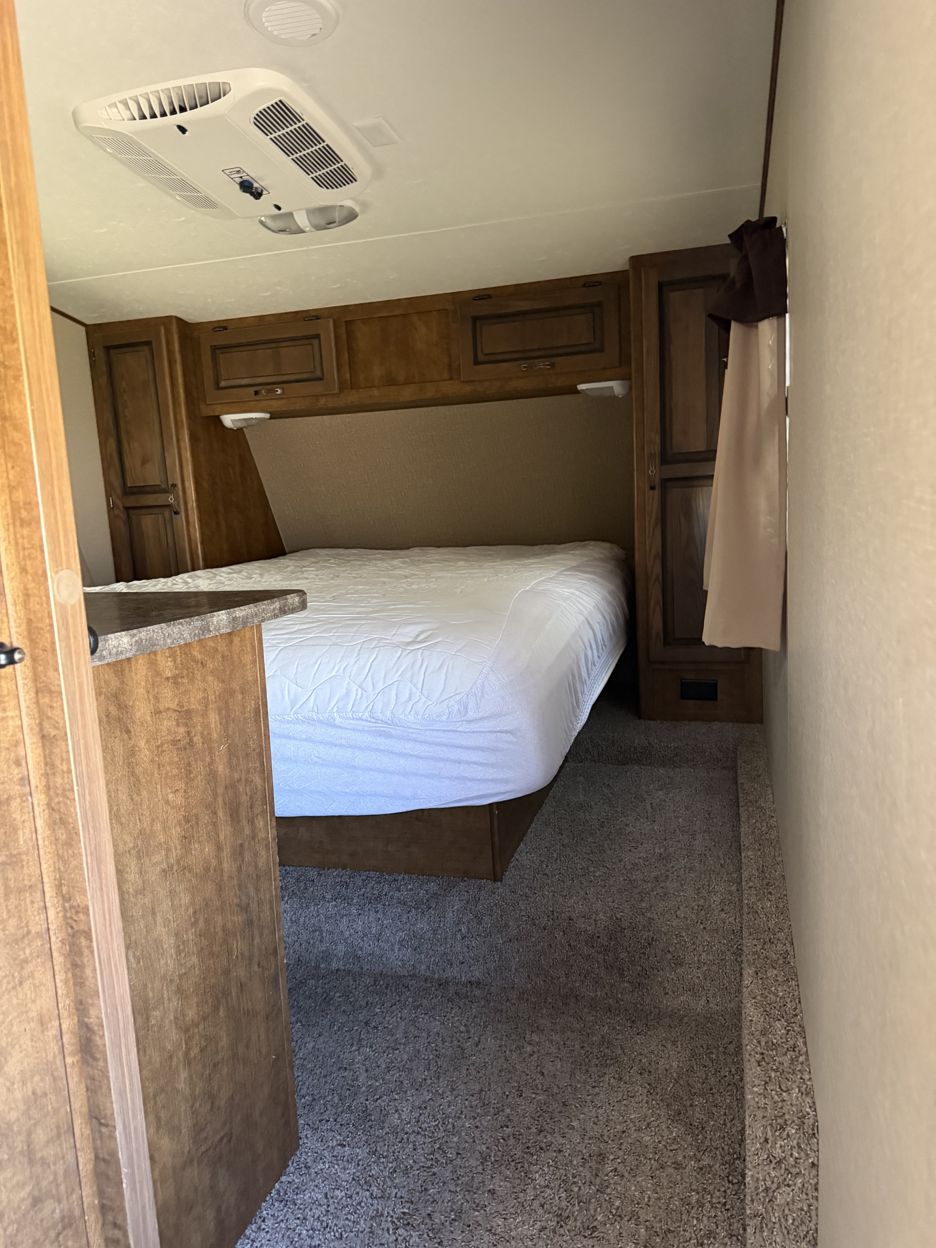 Cozy RV rental bedroom featuring a queen bed with white sheets, wooden cabinets, an overhead AC unit, and carpeted floor.. Crossroads RV Rezerve 2015