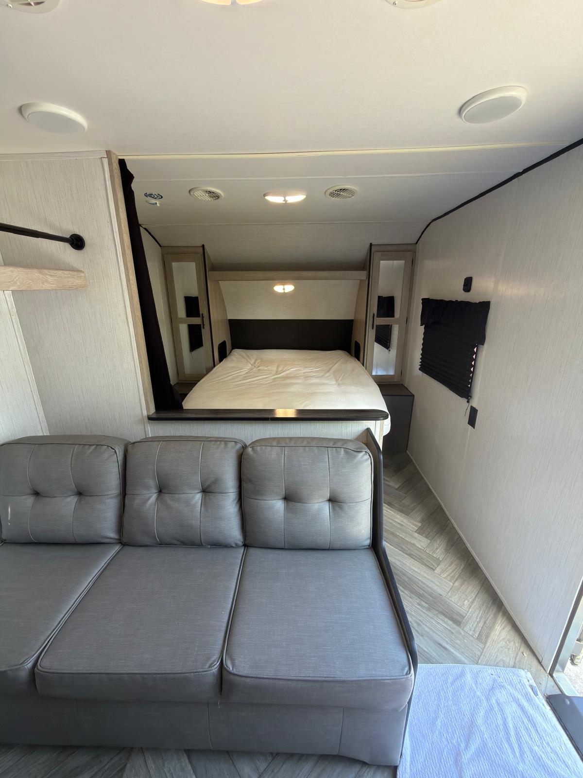 RV rental interior: A cozy gray sofa faces a compact bedroom with a neatly made bed and mirrored bedside cabinets.. Heartland RVs Prowler 2022
