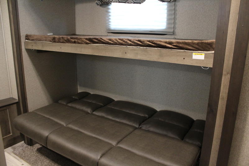 RV rental sleeping area showing an upper bunk bed with a blanket and a lower dark segmented sofa bed for travelers.. Keystone RV Sprinter Campfire Edition 2019