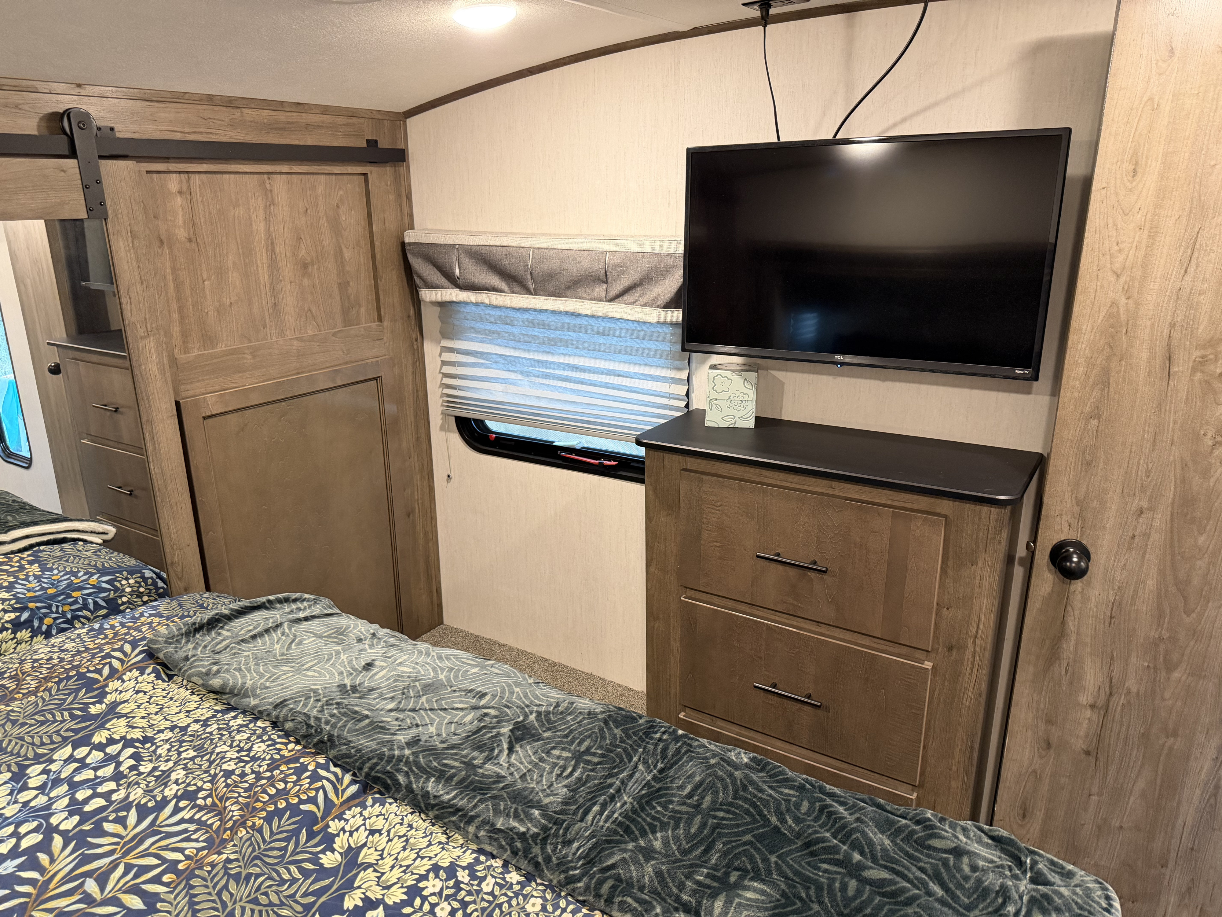 Explore this cozy RV rental bedroom. Features a bed with patterned linens, a mounted TV, window, and wood-toned dresser.. Forest River Sabre 2022