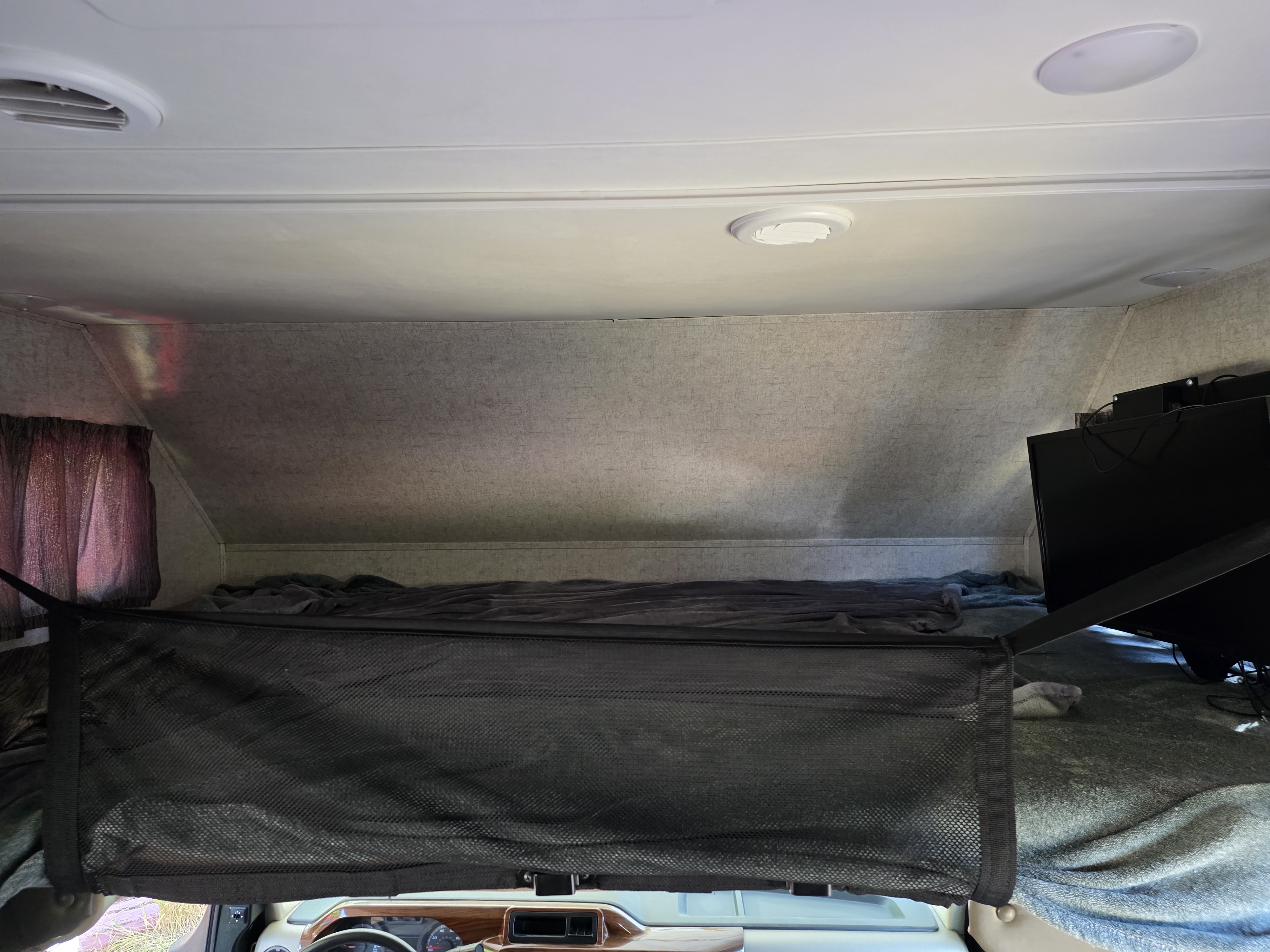 RV rental's cab-over bunk area: a bed with blankets, safety net, ceiling vents/lights, and a TV.. Forest River Forester 2017