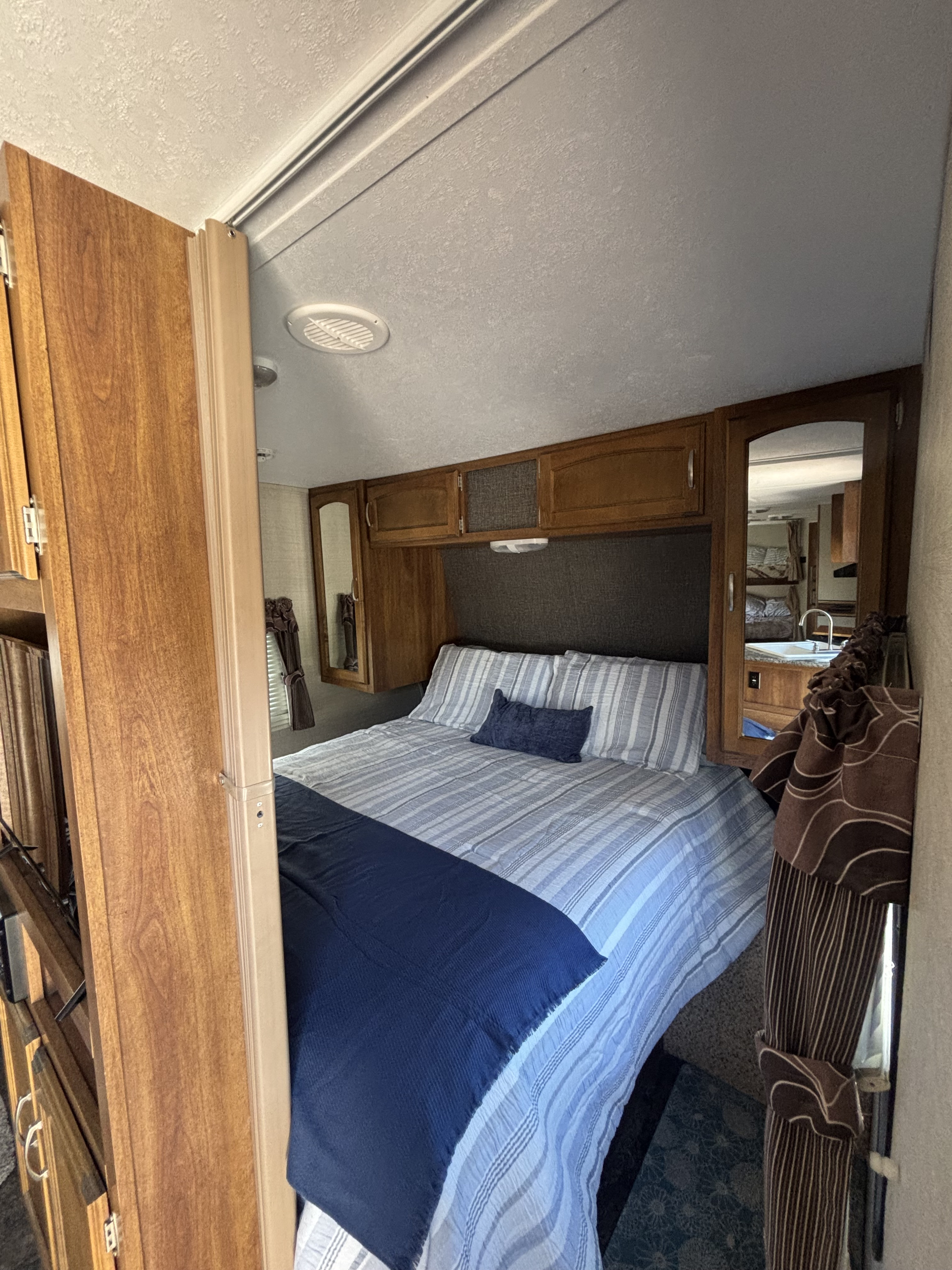 RV rental bedroom showcasing a queen bed with blue striped bedding, wood cabinets, and a mirrored closet reflecting the galley.. Keystone RV Springdale 2015