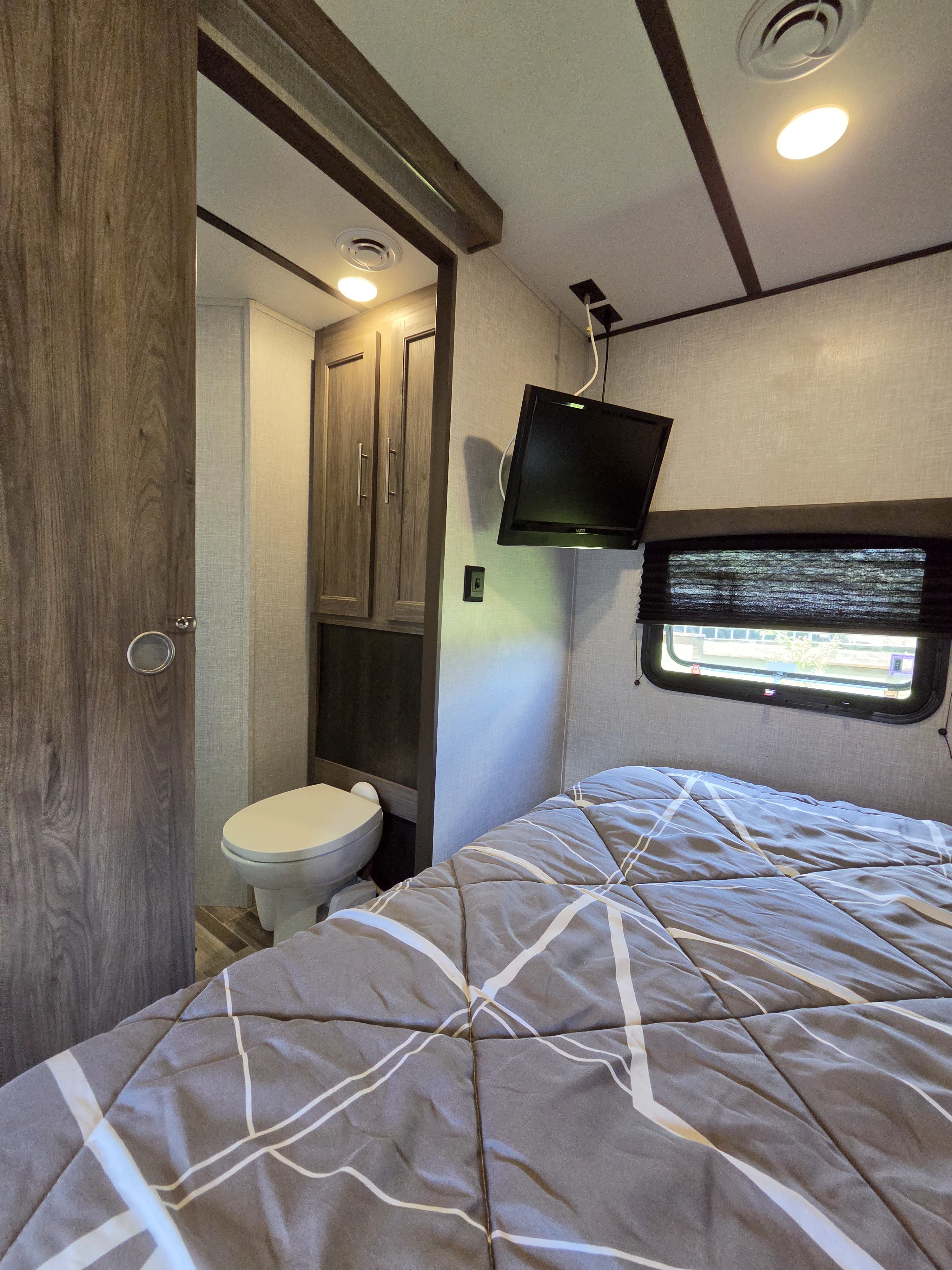 View inside an RV rental showing a cozy bedroom with bed, wall-mounted TV, and a small toilet area for travel convenience.. Crossroads RV Zinger 2021