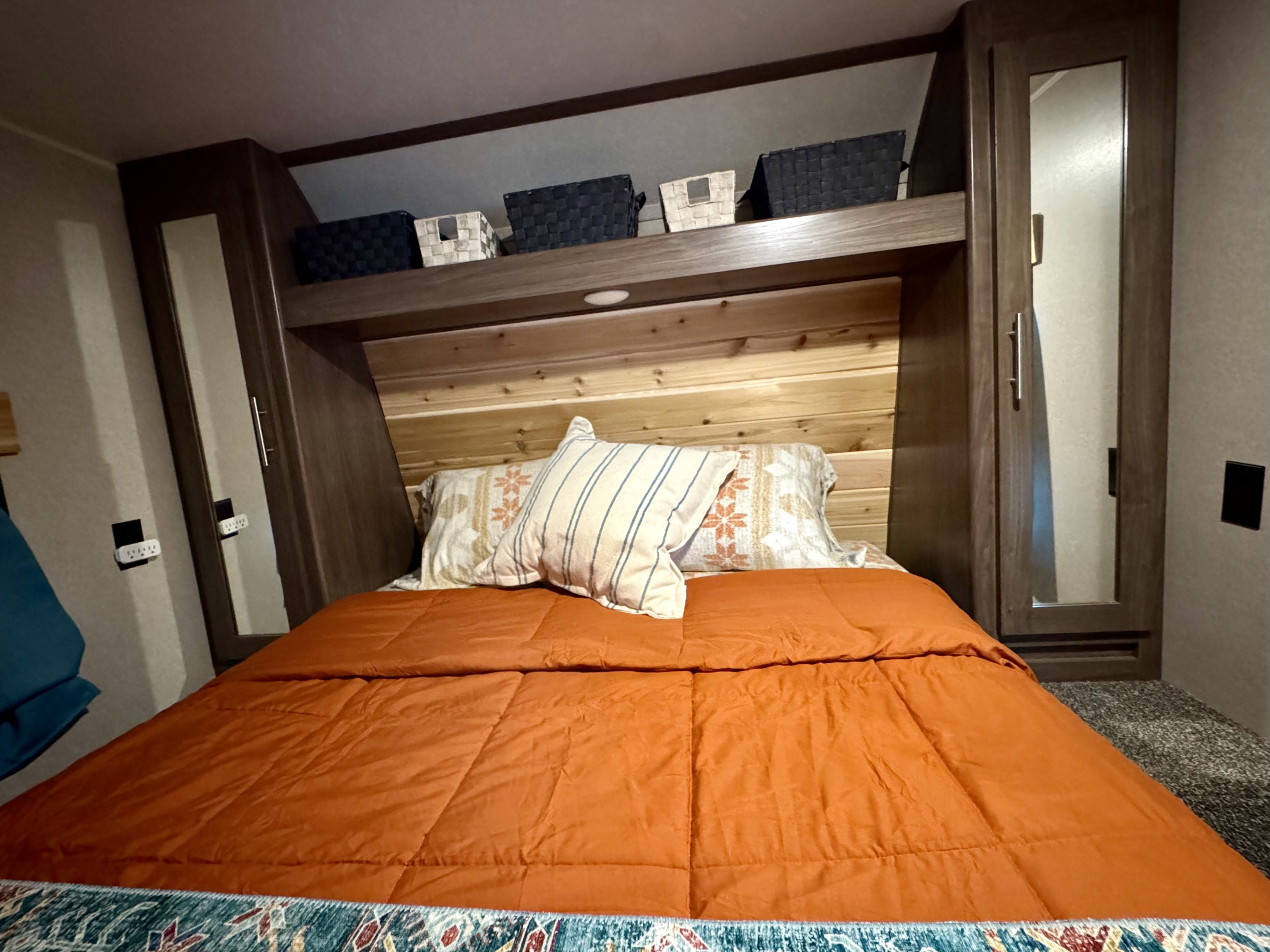 Cozy rv rental bedroom featuring a bed with an orange comforter, patterned pillows, a wooden headboard, overhead storage with baskets, and mirrored cabinets.. Keystone RV Hideout LHS 2019