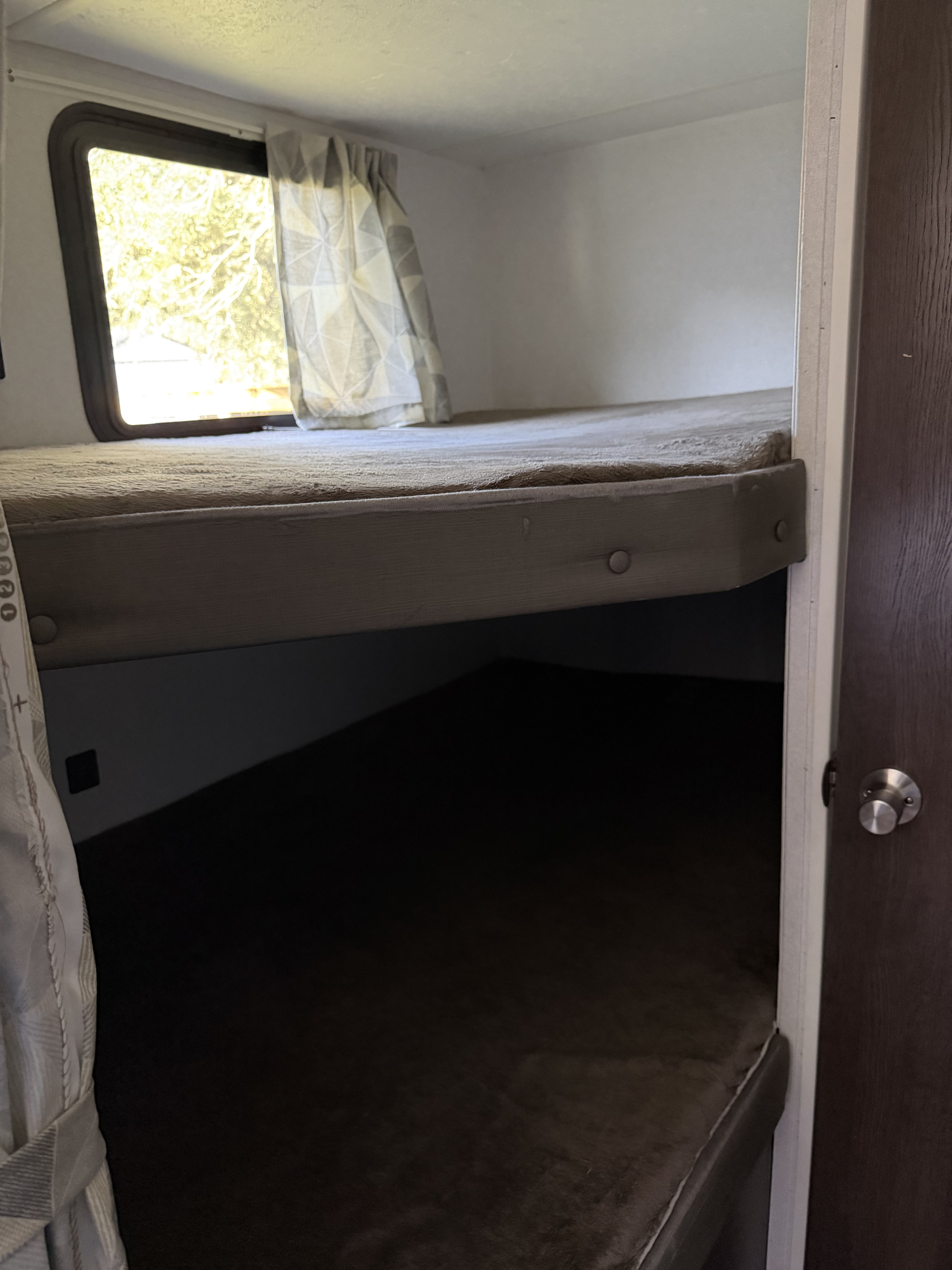 RV rental bunk bed setup: upper bunk with window and curtain, lower bunk in shadow, seen from an adjacent open door.. Forest River Salem Cruise Lite 2019